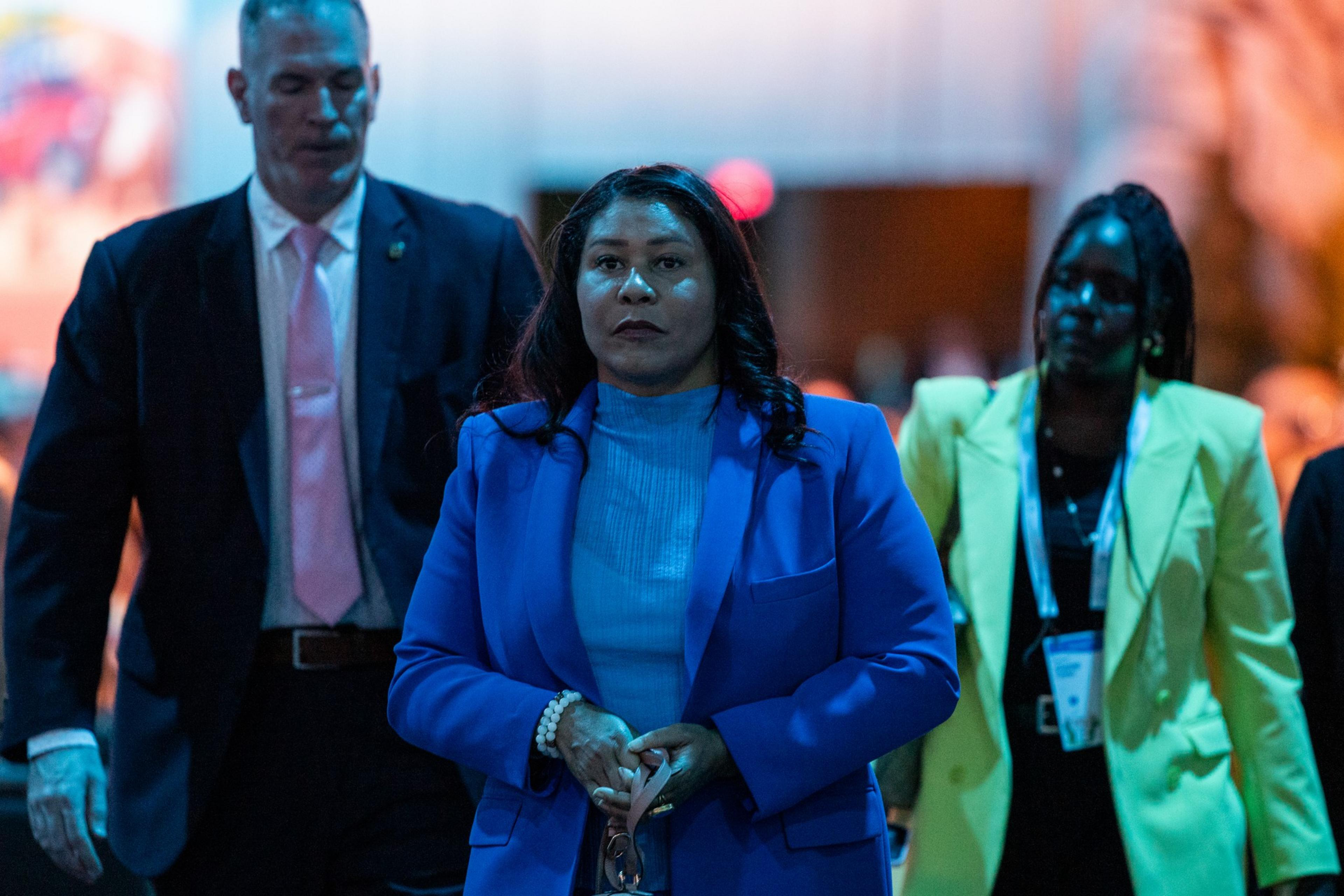 A woman in a blue blazer stands between a man in a dark suit with a pink tie and a woman in a yellow blazer, walking in an indoor setting.