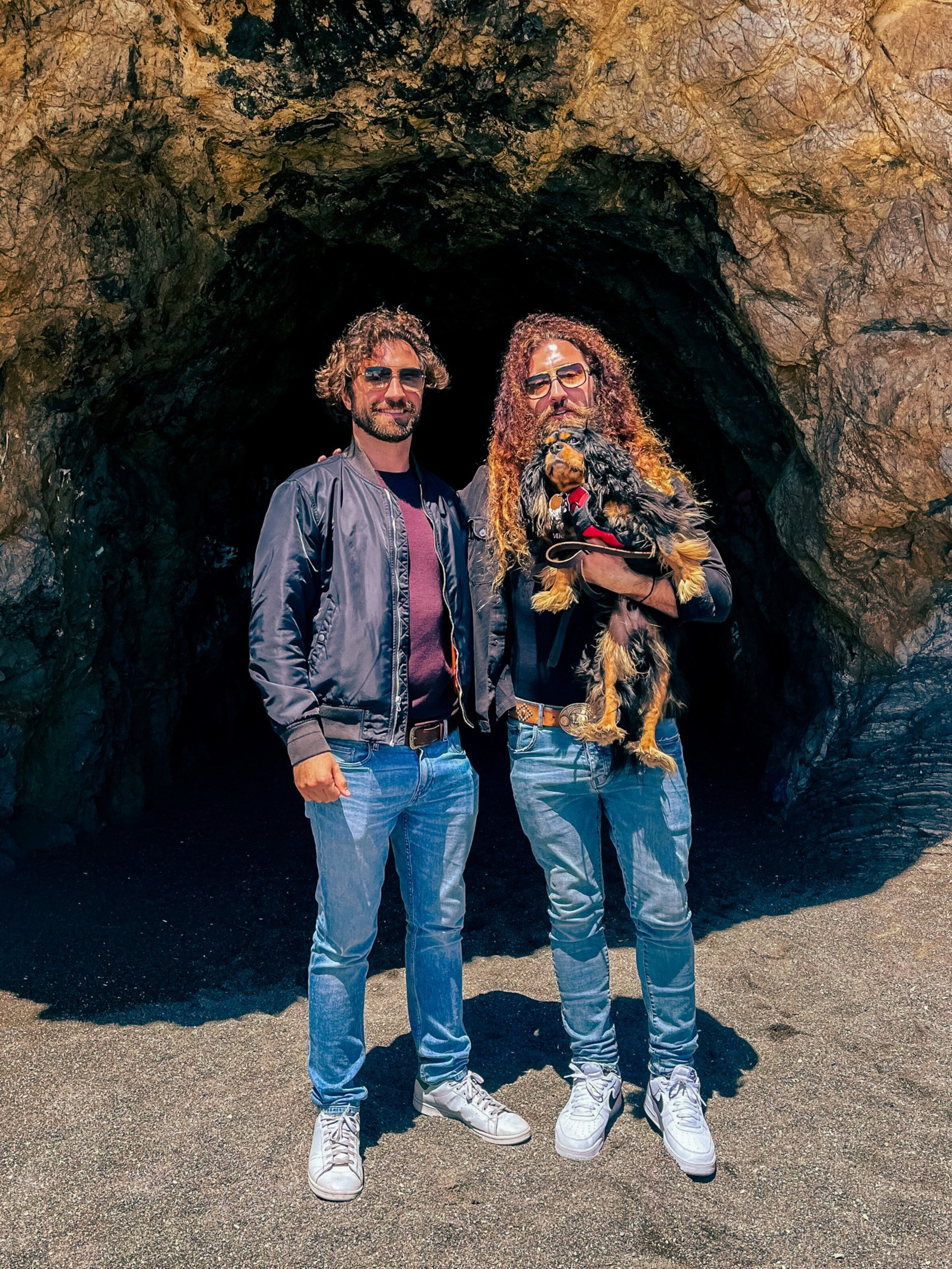 Two people stand in front of a rocky cave entrance, one holding a small dog. They are wearing casual clothes and sunglasses, and the sun casts clear shadows.