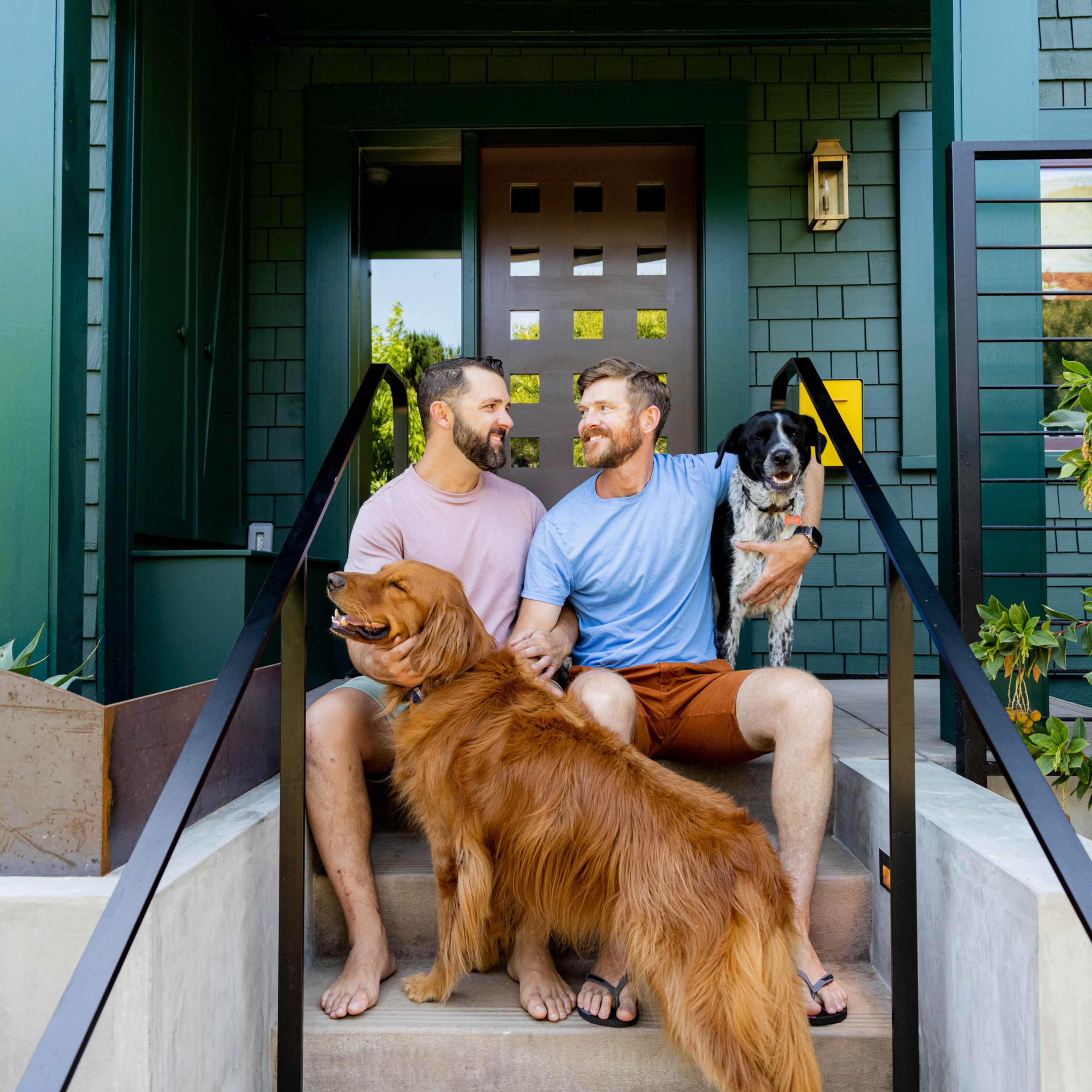 Two men sit on their porch steps, smiling at each other, each petting a dog. One dog is a golden retriever, and the other is a black and white mixed breed.