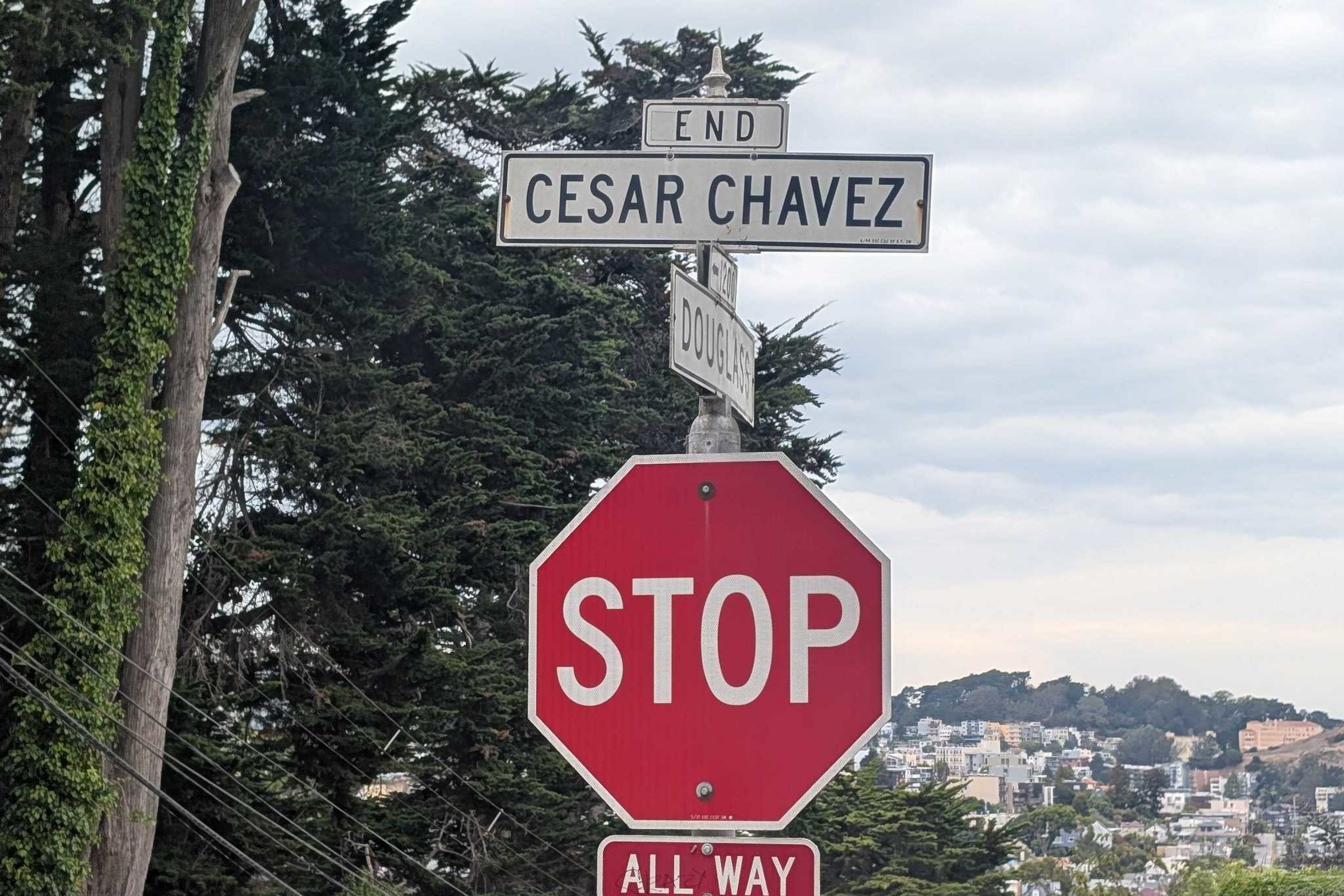 A stop sign with an "ALL WAY" plaque stands below a street sign marking the end of Cesar Chavez street, surrounded by trees and distant buildings.