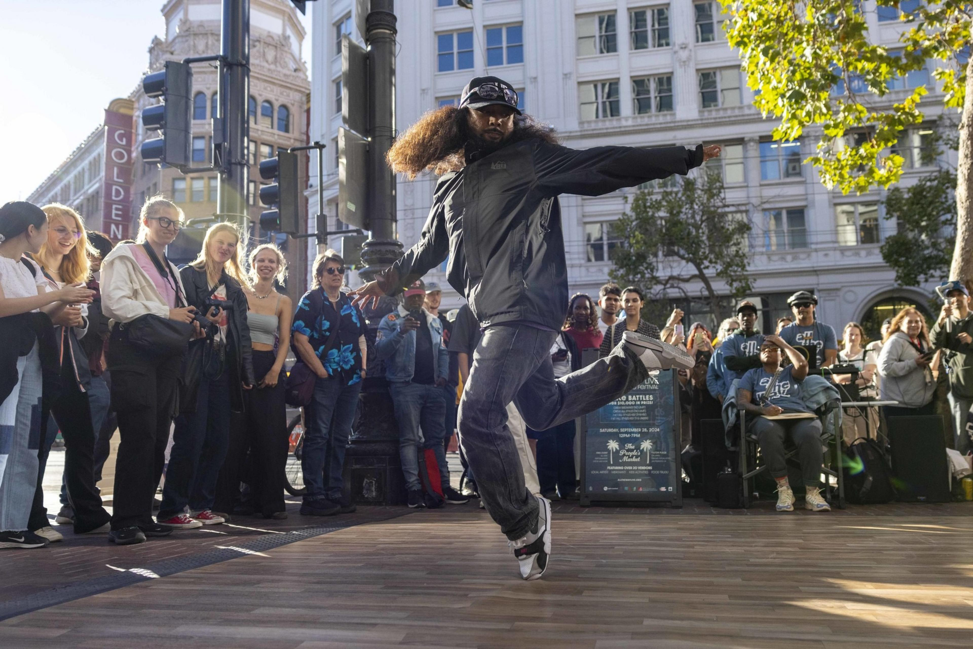 A man with long hair and a cap performs a dance move on a wooden platform in front of an engaged crowd on a city street, with buildings and trees in the background.