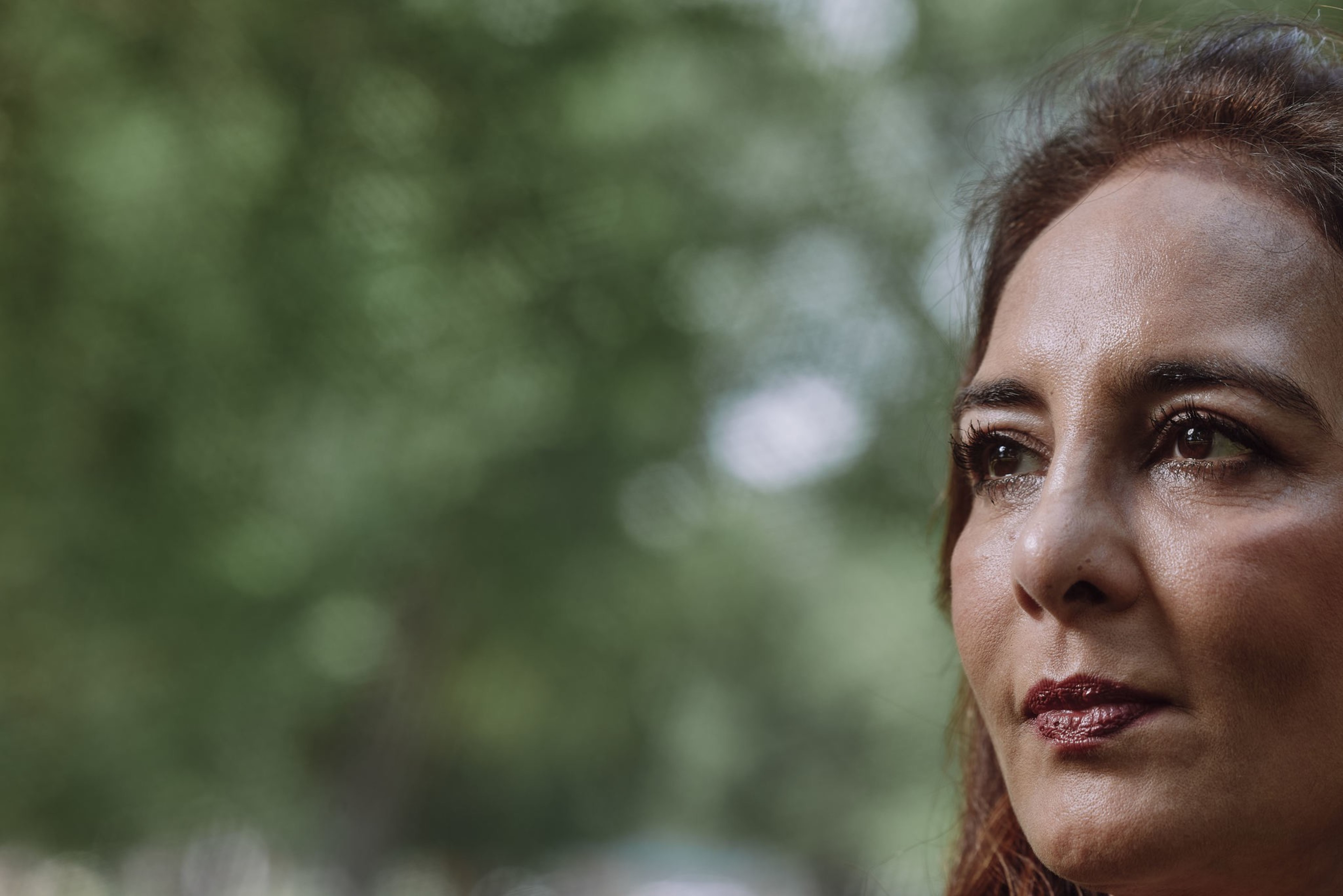 A woman with a thoughtful expression looks into the distance. Her face is partially cropped and the background is a blurred green, indicating foliage.