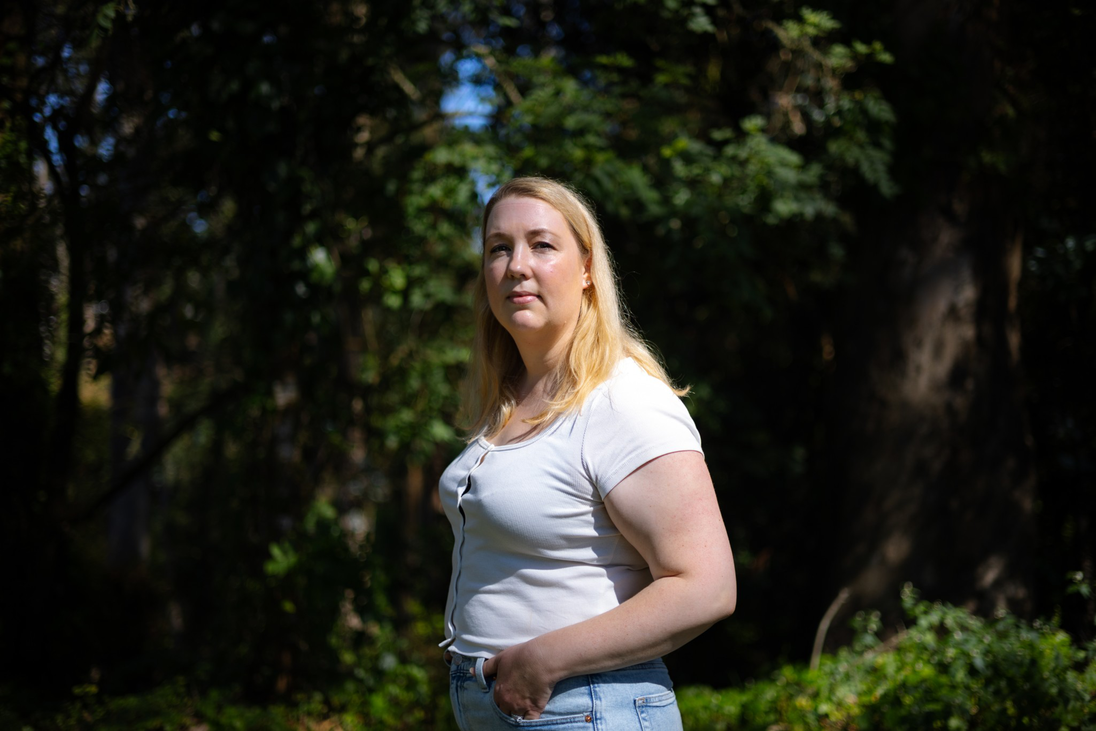 A woman with long blonde hair stands outdoors in a white shirt and blue jeans, with a background of trees and greenery in soft sunlight.
