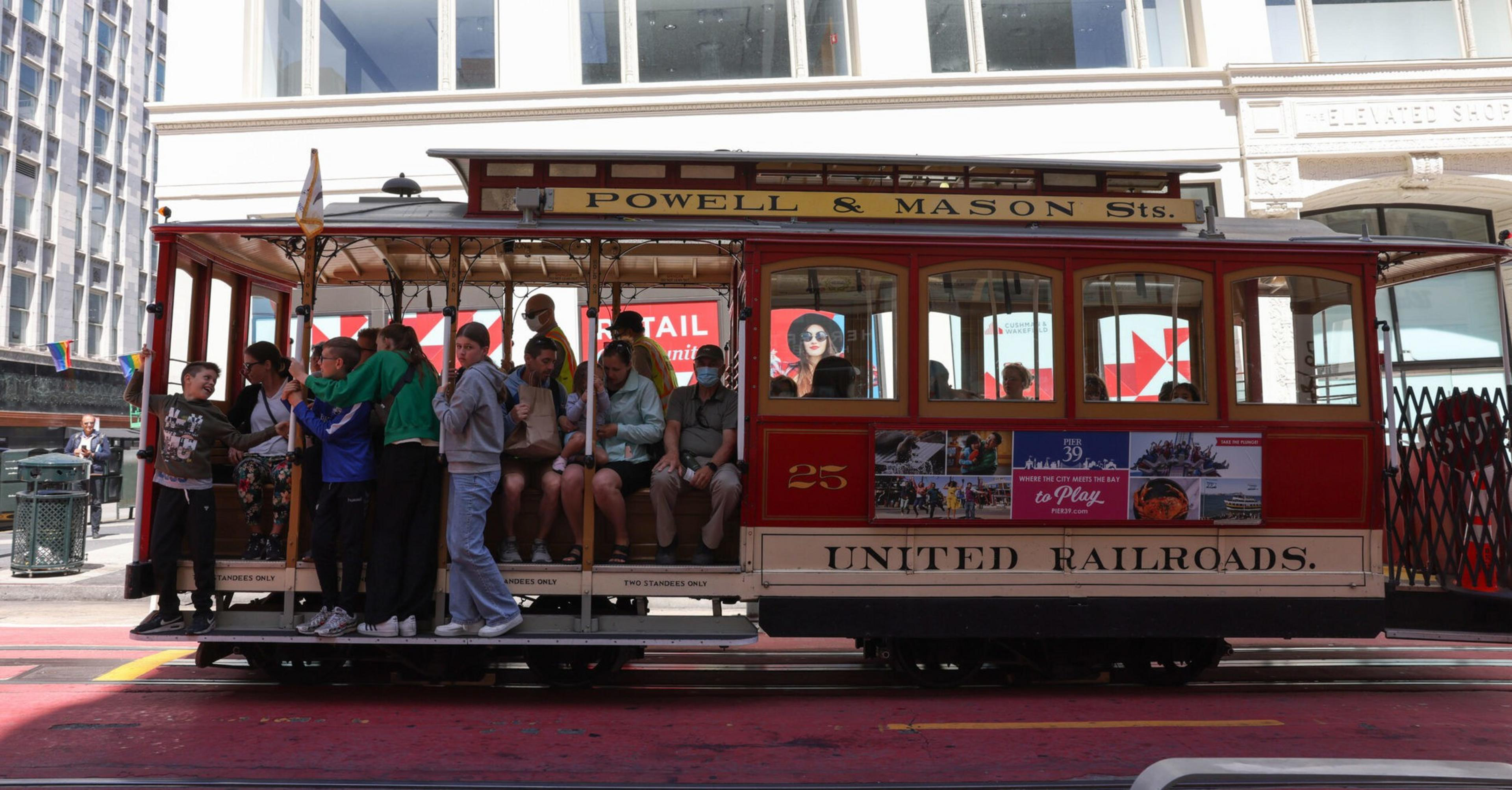 A side view of a red cable car shows passengers stranding and sitting on it.