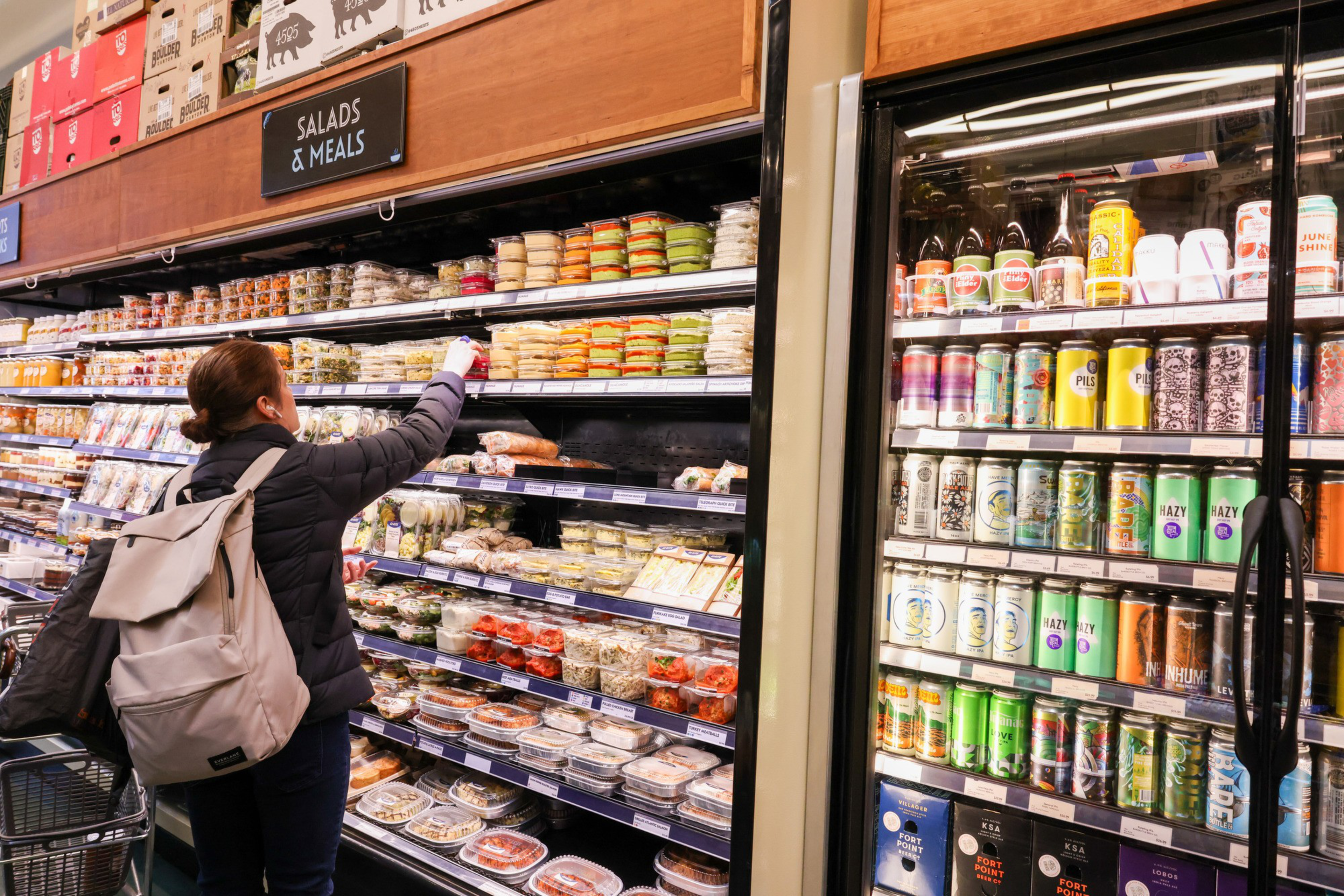 A person with a backpack selects an item from a refrigerated shelf stocked with salads and meals, while cans of beverages fill the adjacent cooler.