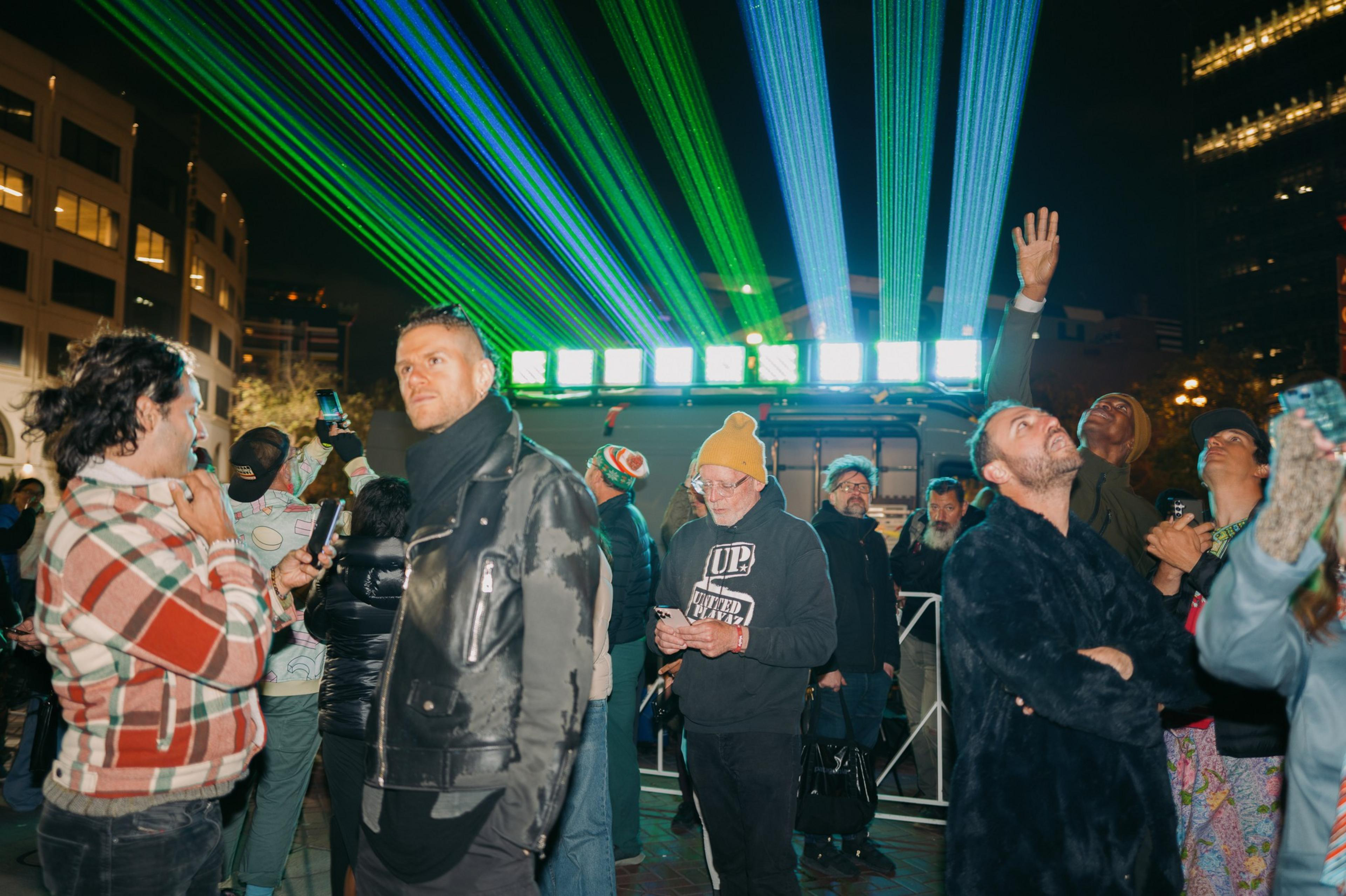 A crowd watches colorful green and blue laser lights in a night scene. People are bundled in jackets and hats, some capturing the moment with phones.