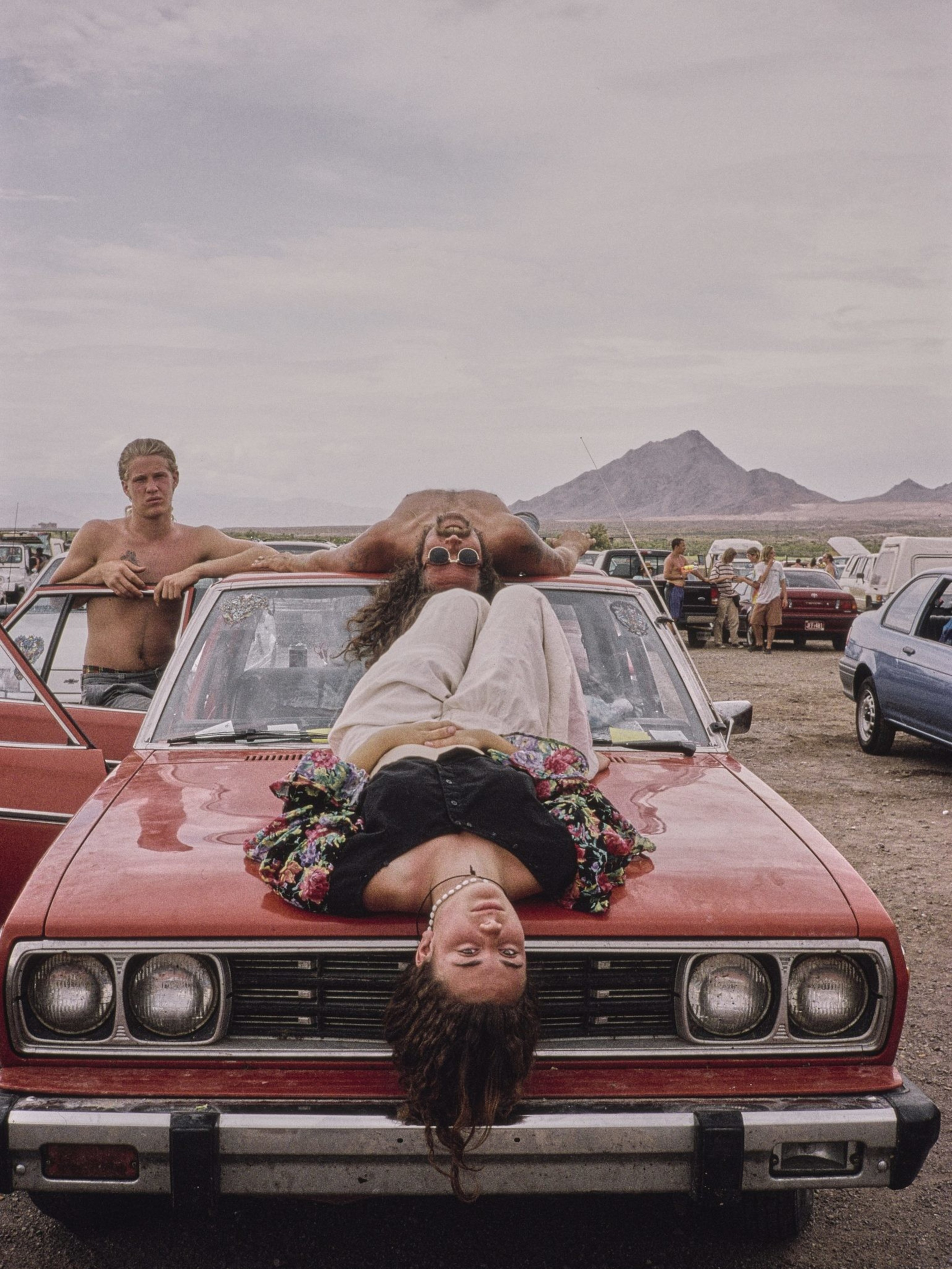 A person is lying on the hood of a red car, relaxed, with another sitting on the windshield and a third standing beside them. Mountains and other cars are in the background.