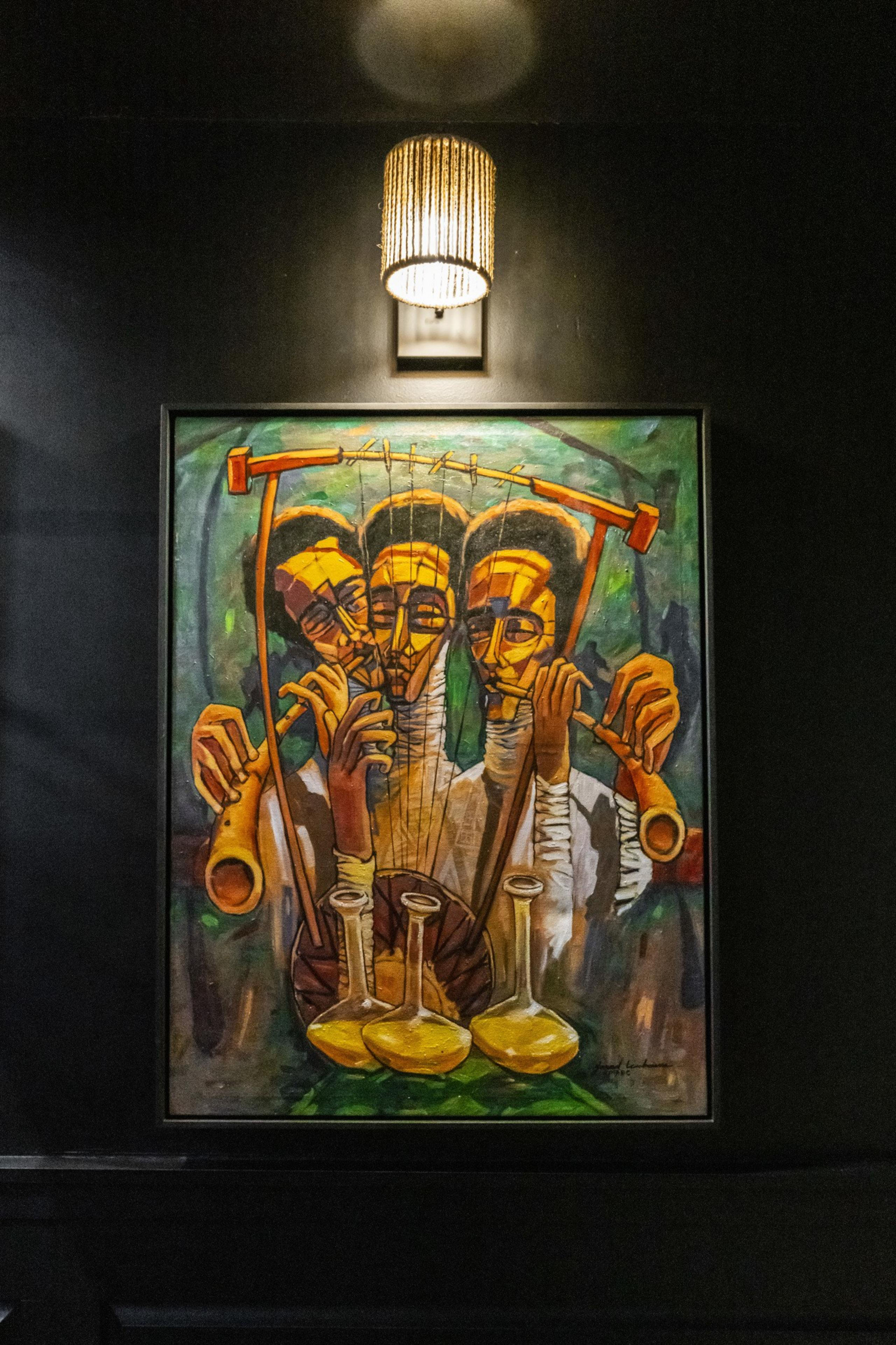 The image shows a colorful painting of three abstract figures playing musical instruments, under a warm, lit sconce on a dark wall background.