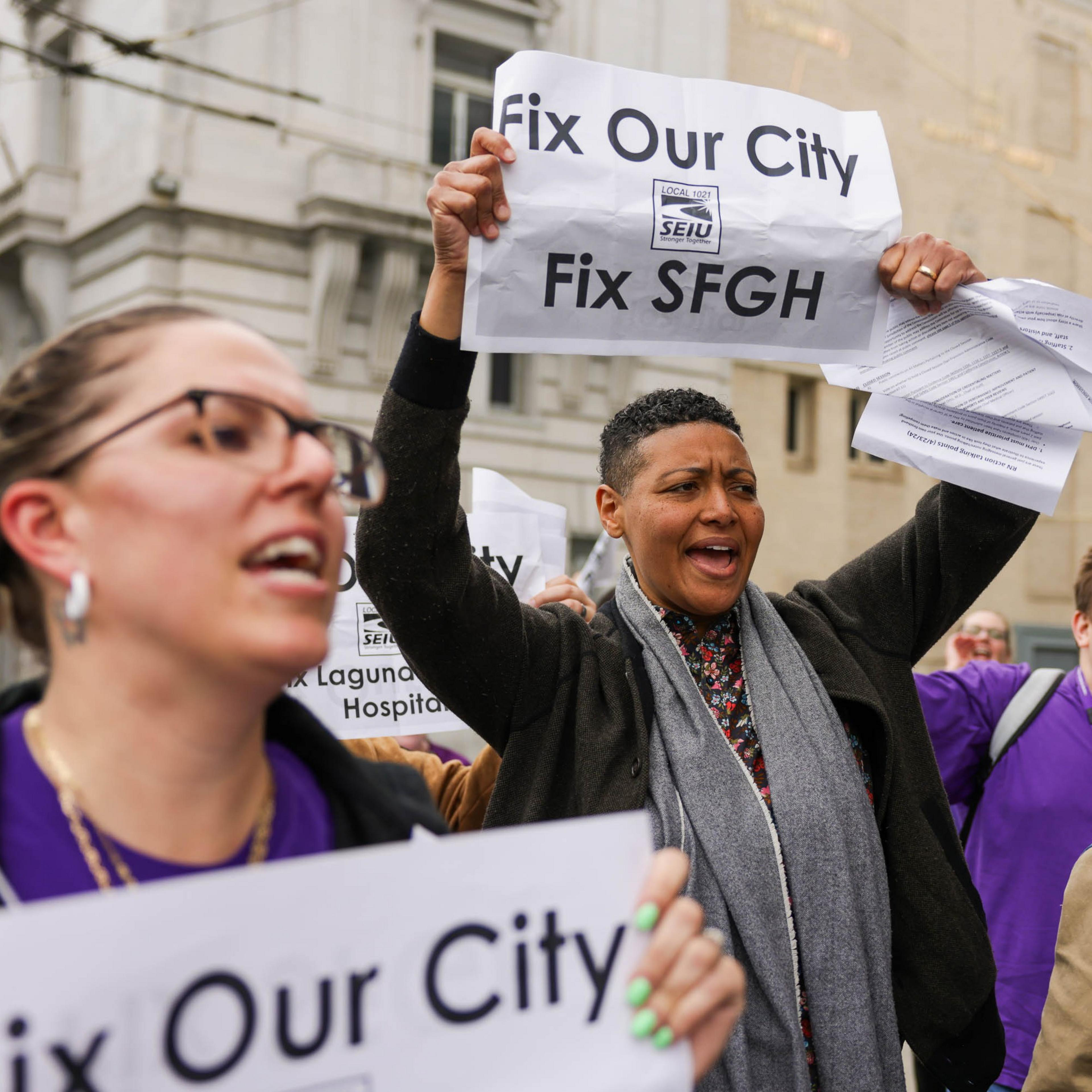 A group of people is protesting outdoors, holding signs reading "Fix Our City" and "Fix SFGH," with determined expressions and wearing purple clothing.