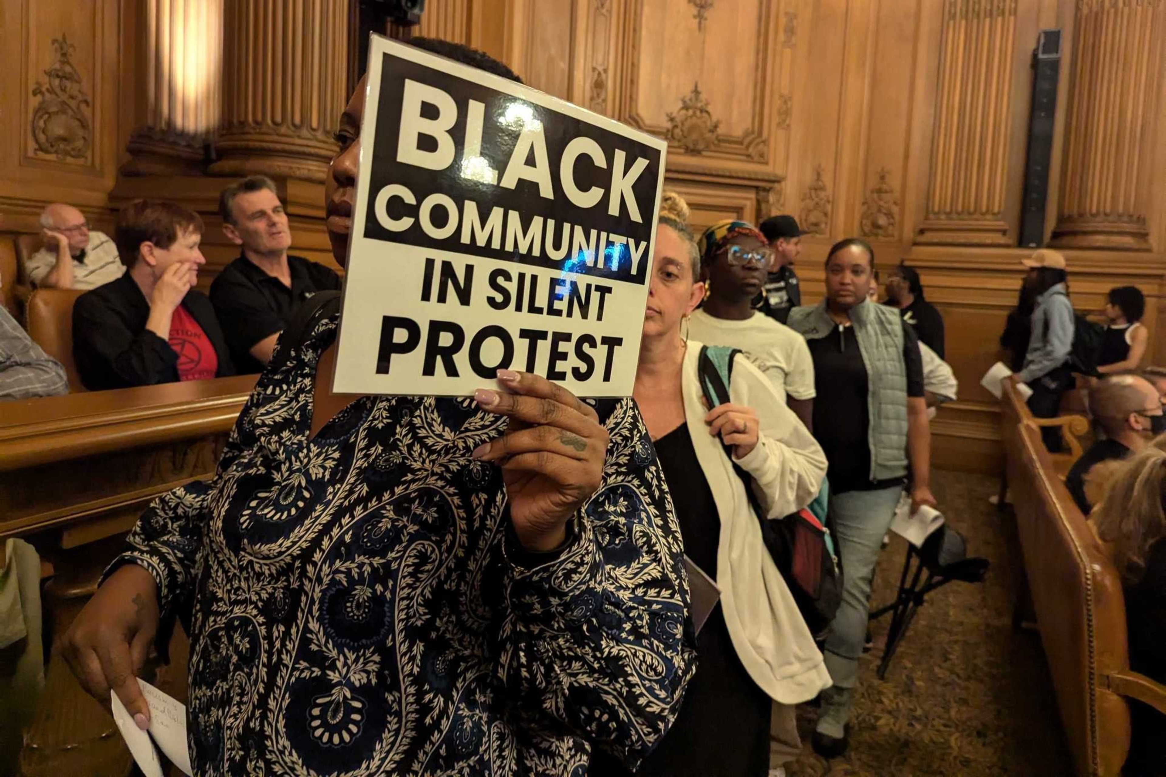 A person holds a sign reading "Black Community in Silent Protest" in a wooden-paneled room filled with people, suggesting a gathering or meeting.