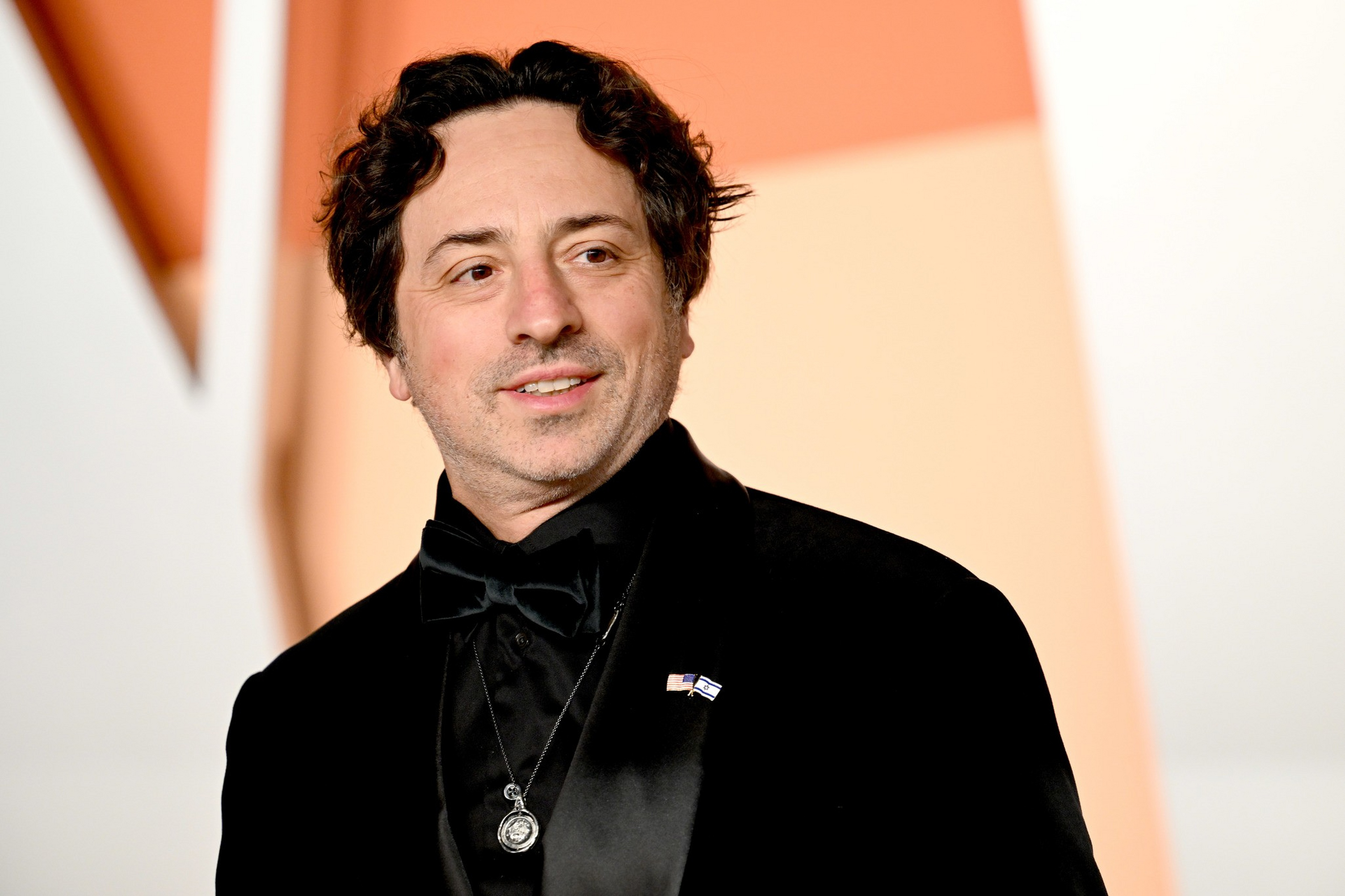 A man with wavy dark hair wearing a black tuxedo, bow tie, and pendant necklace smiles slightly against a warm-toned background.