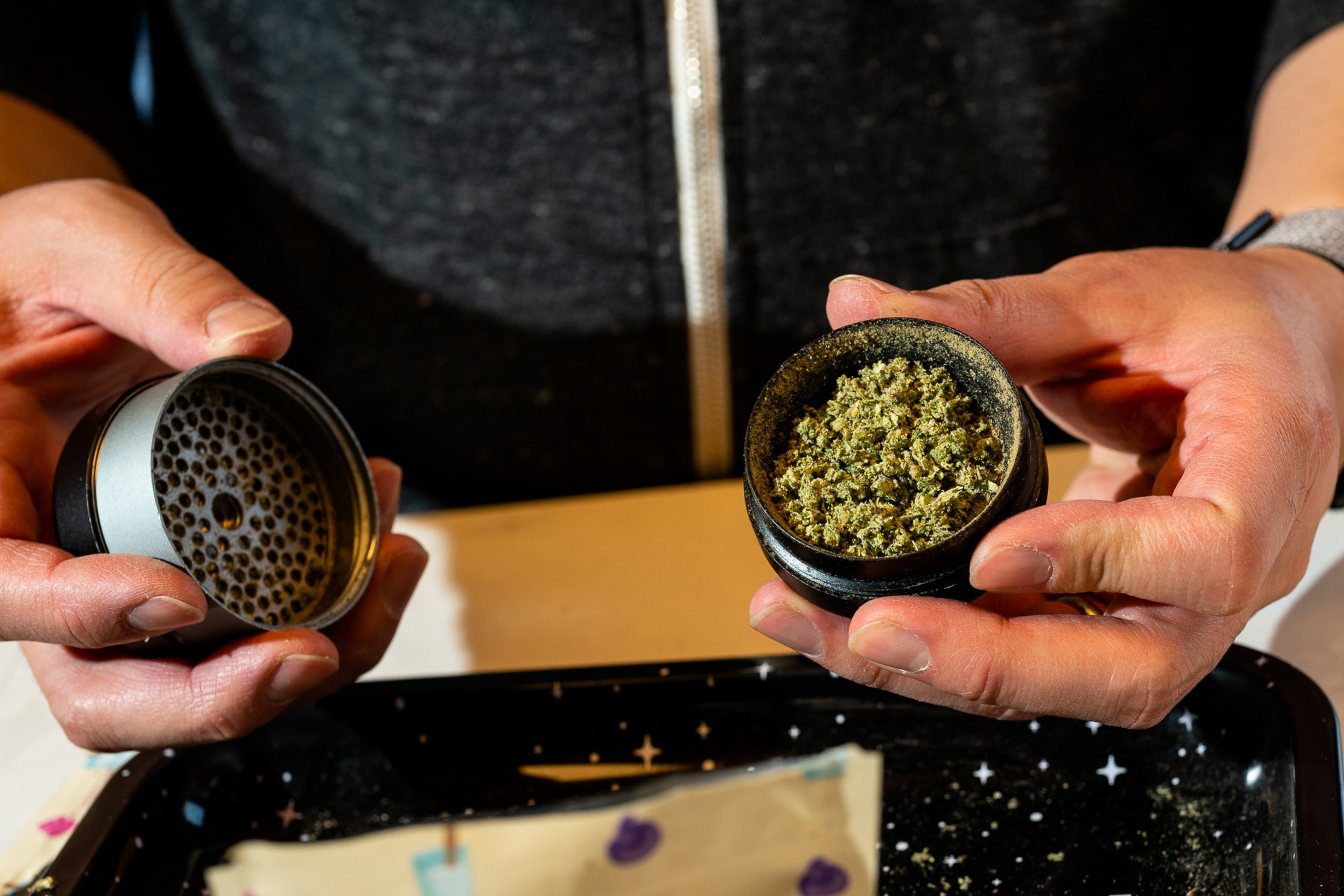 A person holds a two-piece grinder with ground cannabis inside, over a tray with star patterns and a package. They appear to be preparing cannabis.
