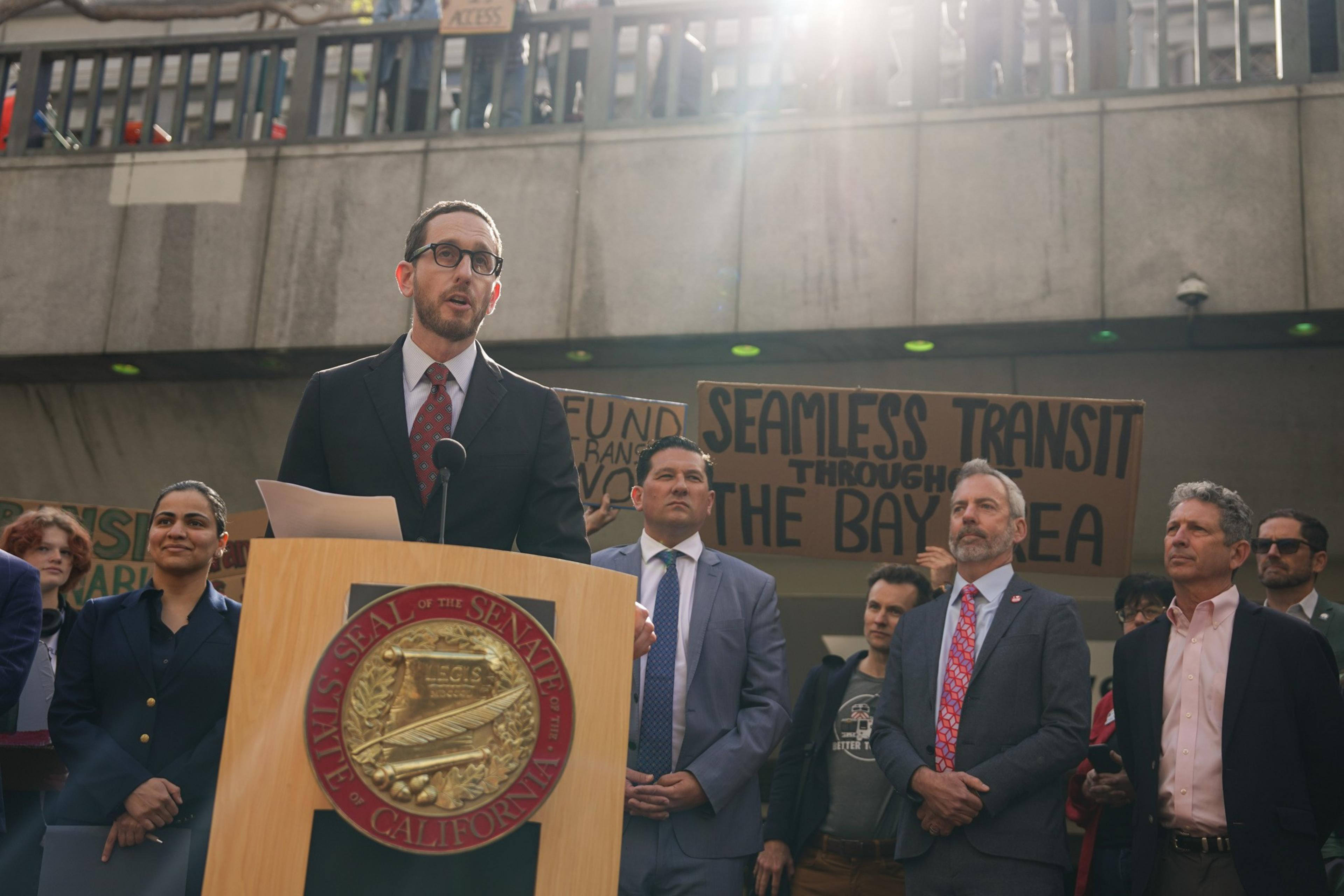 A man speaks at a podium with the California Senate seal; listeners and a protest sign are behind him.