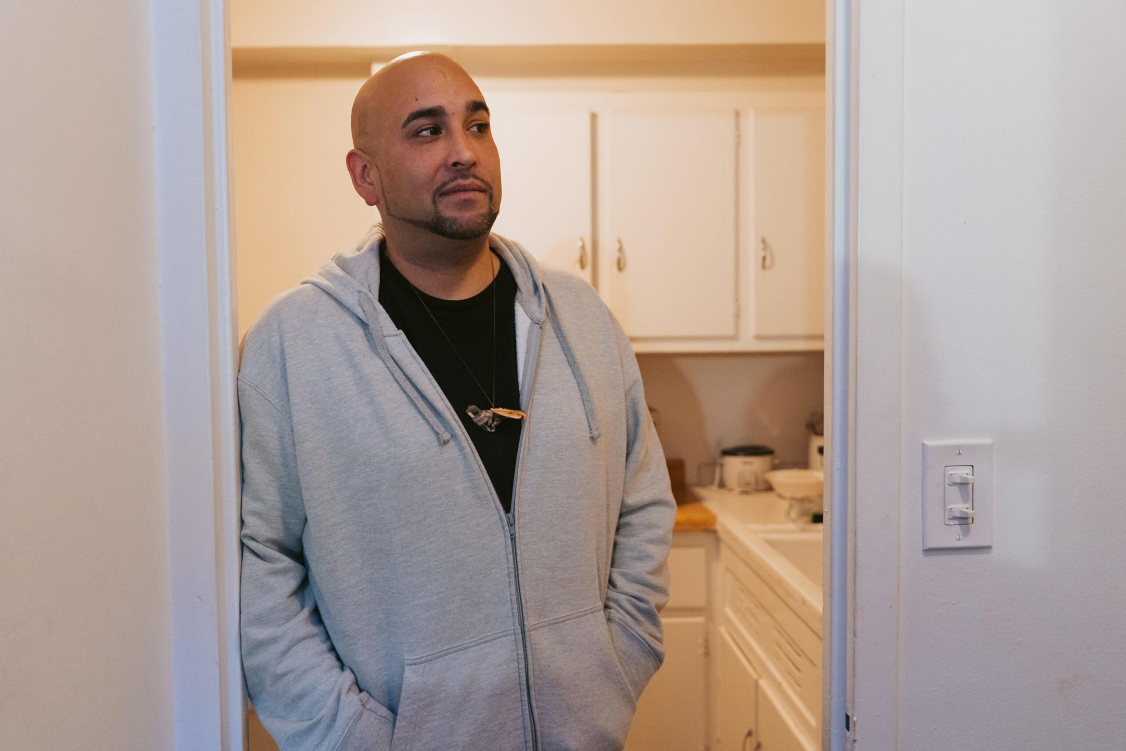 A man with a shaved head and beard, wearing a gray hoodie and black shirt, stands in a doorway looking thoughtfully to the side.
