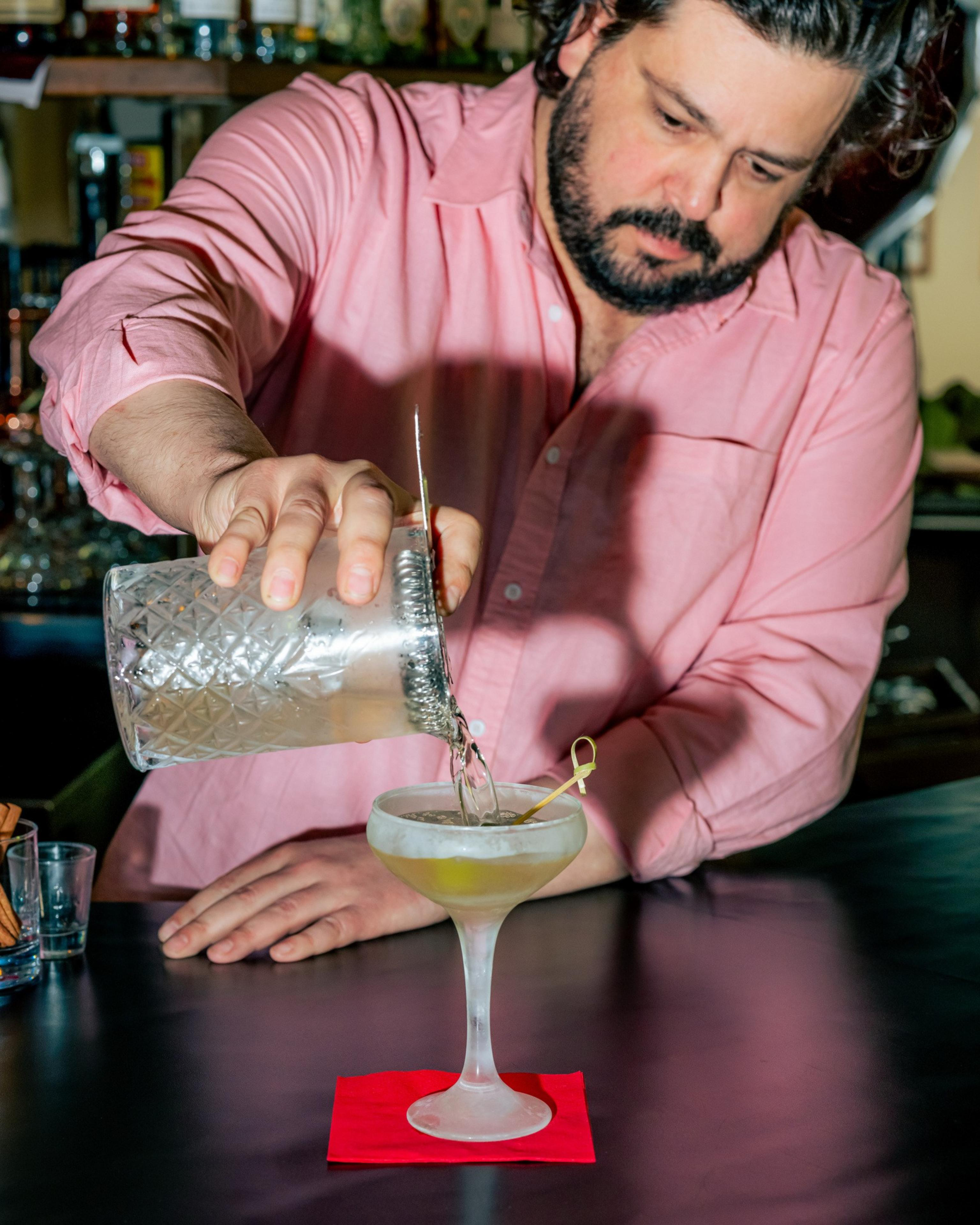 A bartender in a pink shirt pours a clear drink into a frosted cocktail glass on a red napkin. The glass contains a green olive garnish.