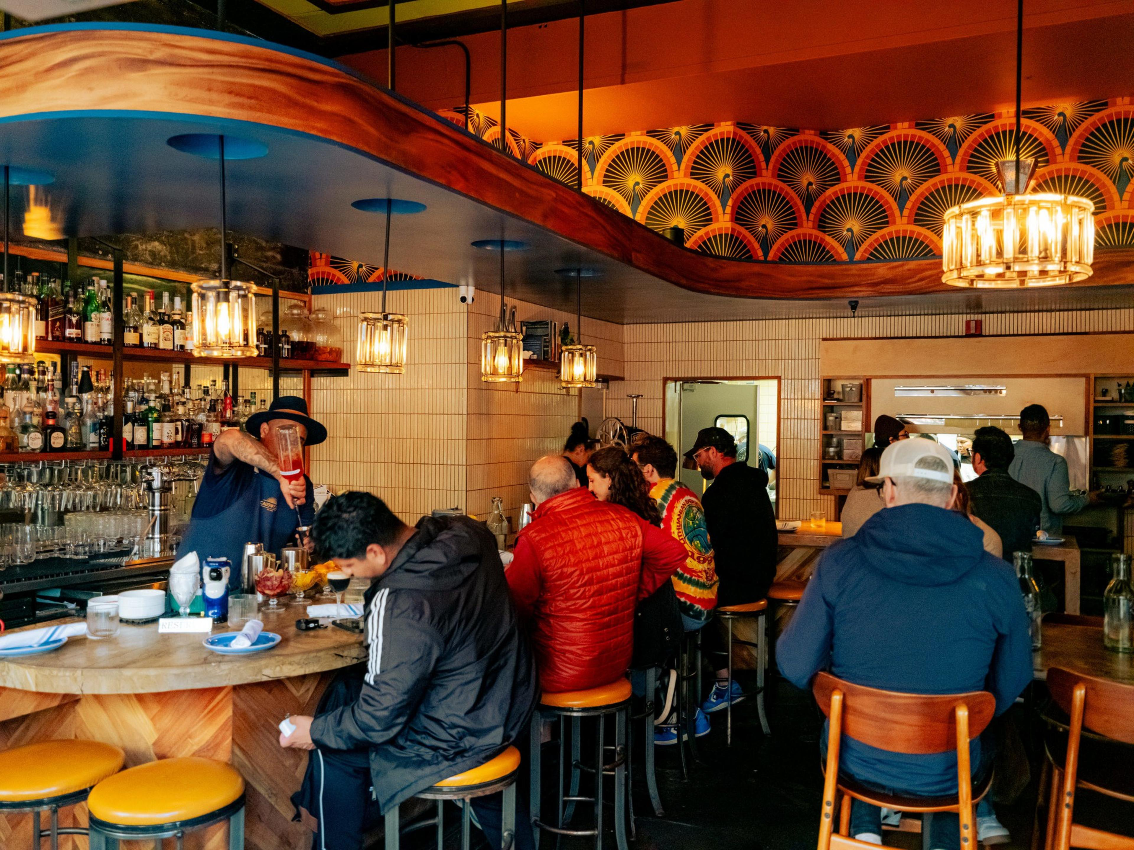 A lively bar scene with a bartender preparing drinks. Patrons sit at a curved bar with colorful stools. The decor features vibrant patterns and hanging lights.