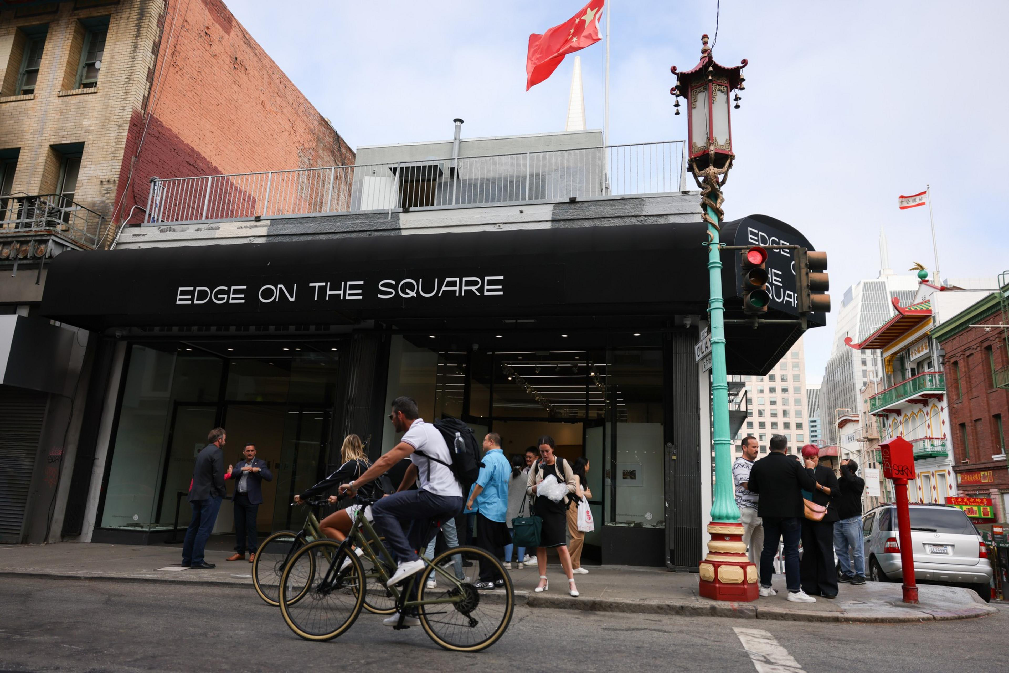 A black awning reading "EDGE ON THE SQUARE" covers a storefront with pedestrians gathered outside and a cyclist riding past on the street.