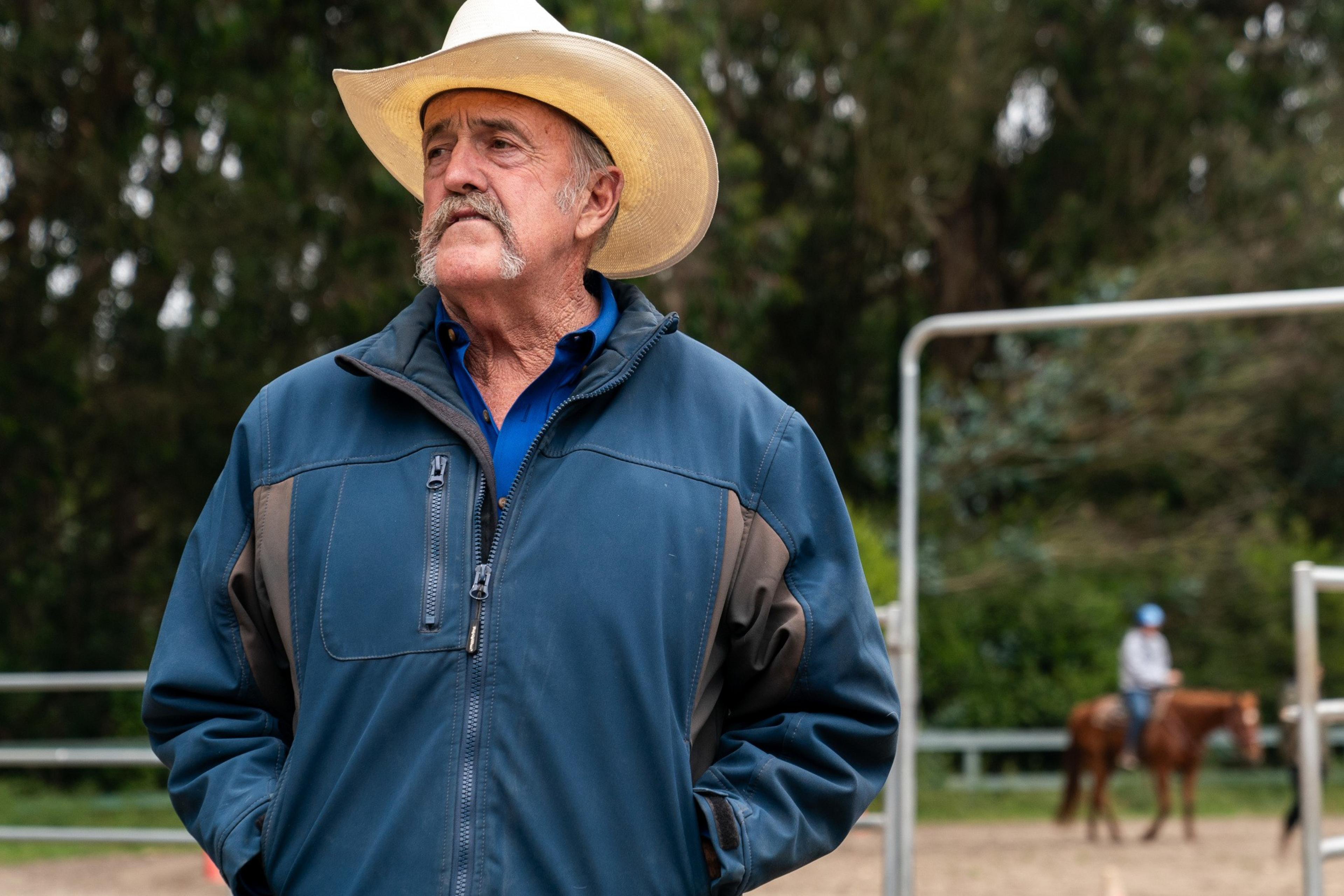 A senior man in a cowboy hat and blue jacket stands outdoors, looking pensive. In the blurred background, a person rides a horse.