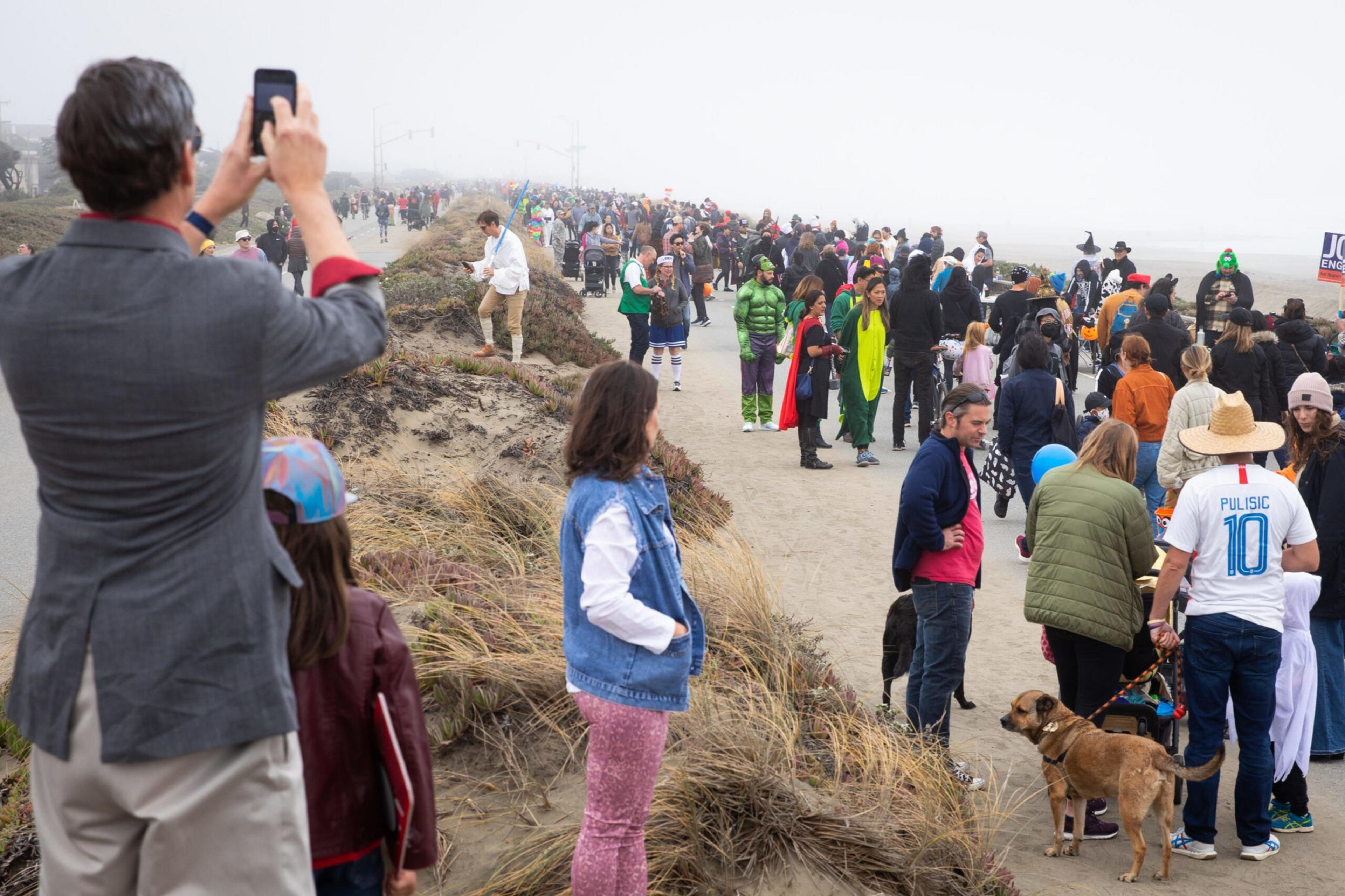 A large crowd of people, some in costumes, walk along a beach path on a foggy day, with a few dogs and spectators watching from the side.
