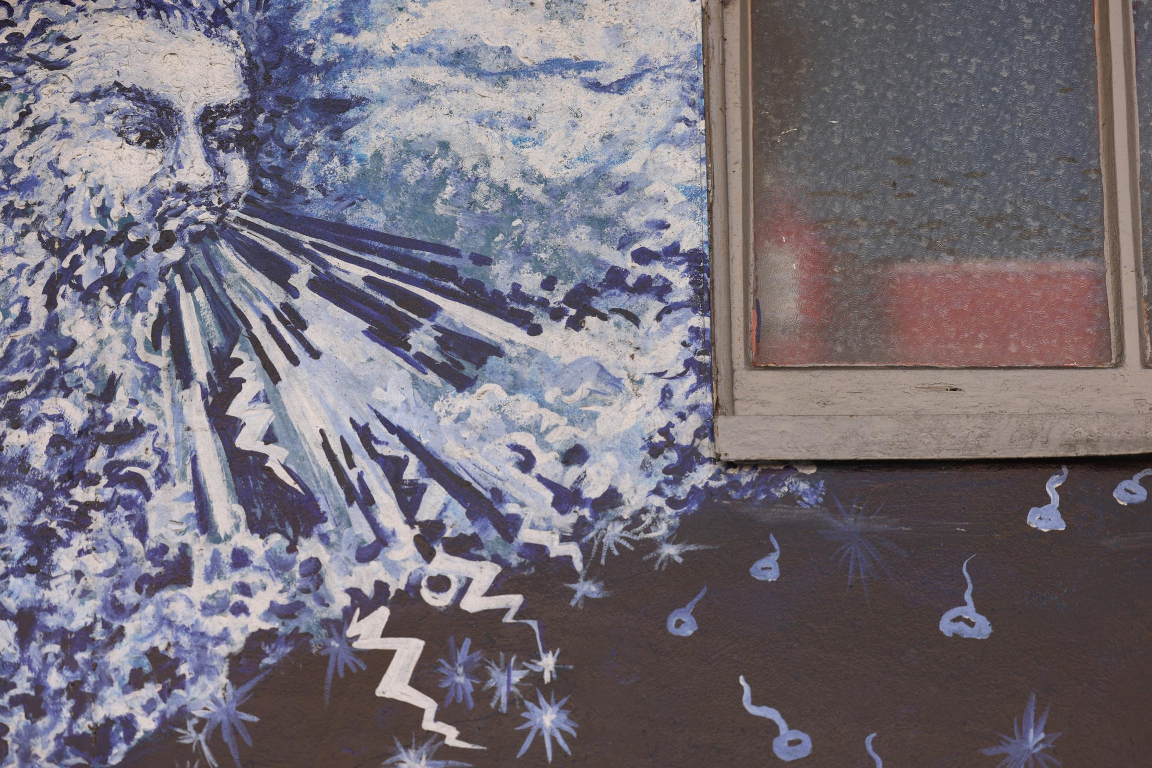 A blue and white mural depicts a face with closed eyes blowing wind or energy, with zigzag lines and star-like shapes spreading outward near a window.