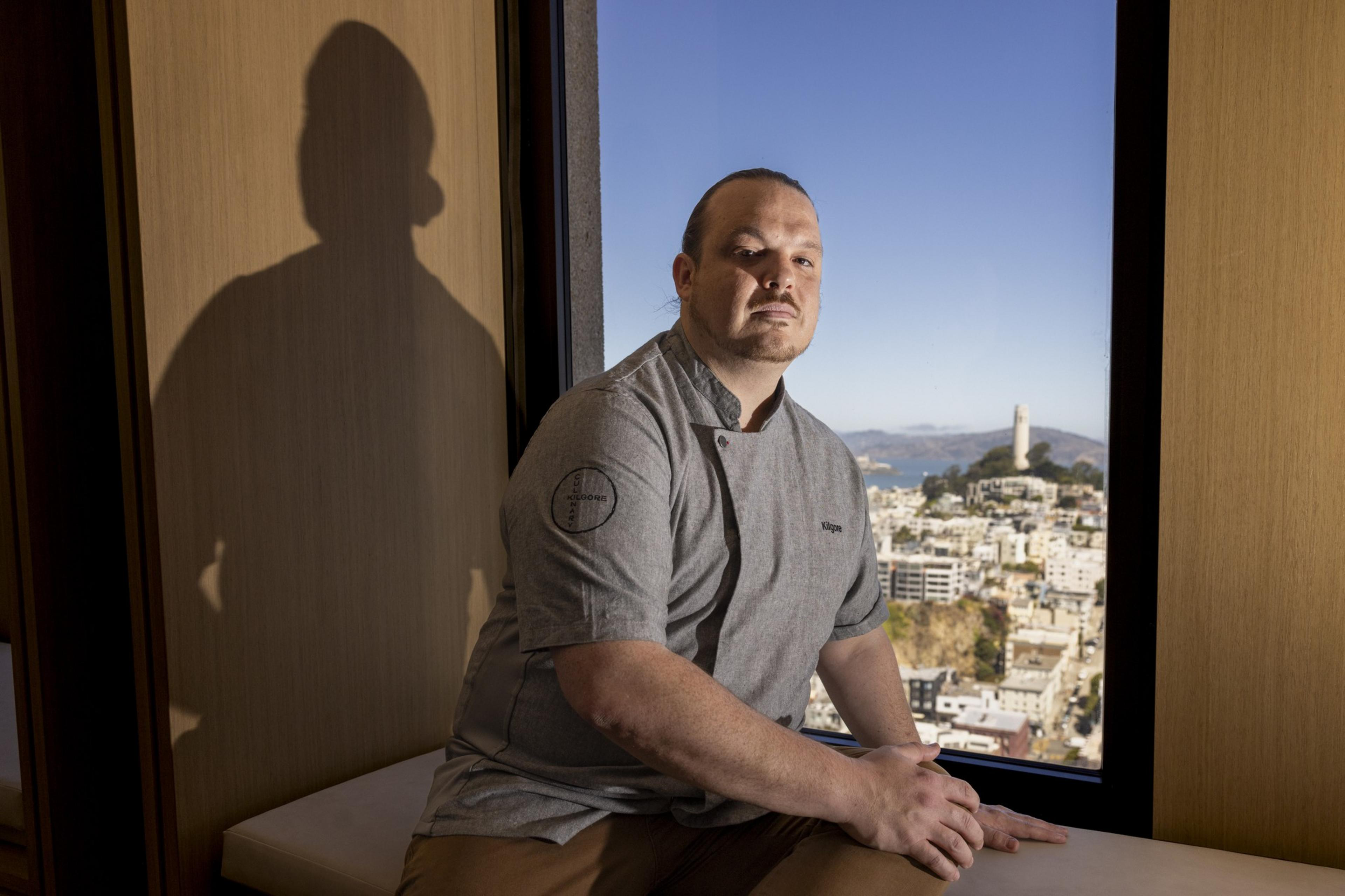 A man wearing a gray chef's coat sits by a window, with a cityscape and a prominent tower in the background. His shadow is cast on a wooden wall beside him.
