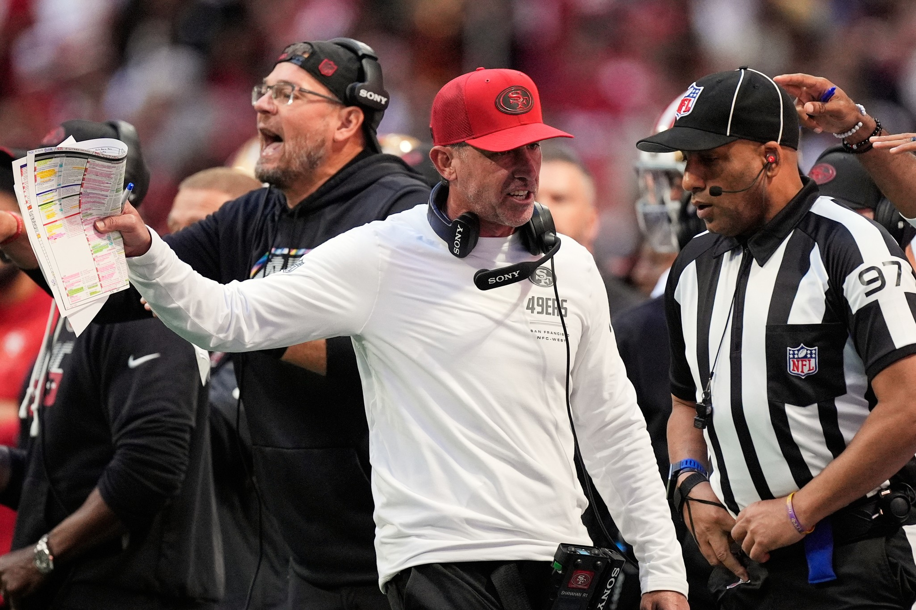 A San Francisco 49ers coach angrily argues with an NFL referee during a game, holding a play sheet in one hand and wearing a red cap.