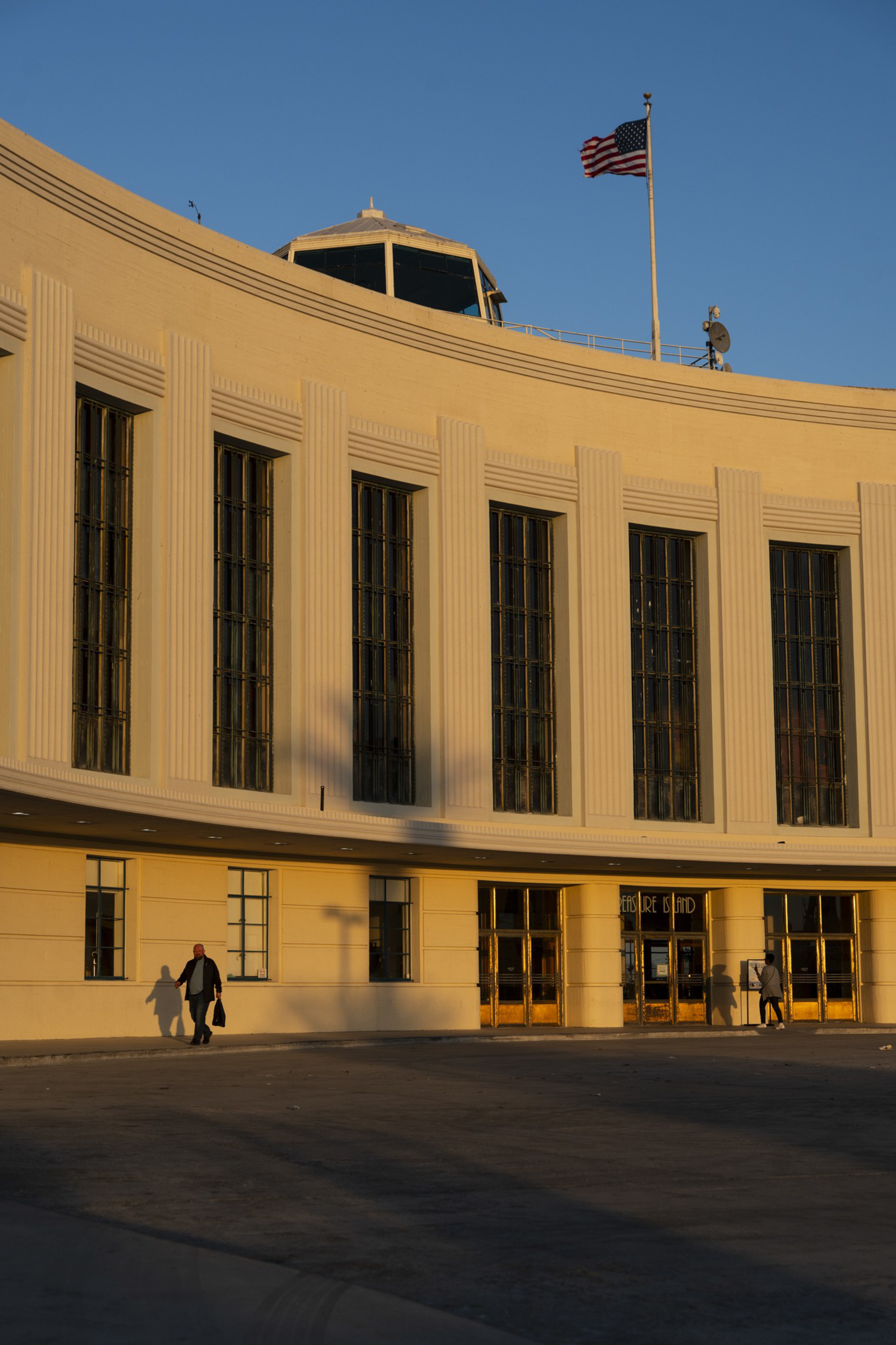 A curved beige building with tall windows and an American flag on top, lit by warm sunlight, with three people walking near its entrances.