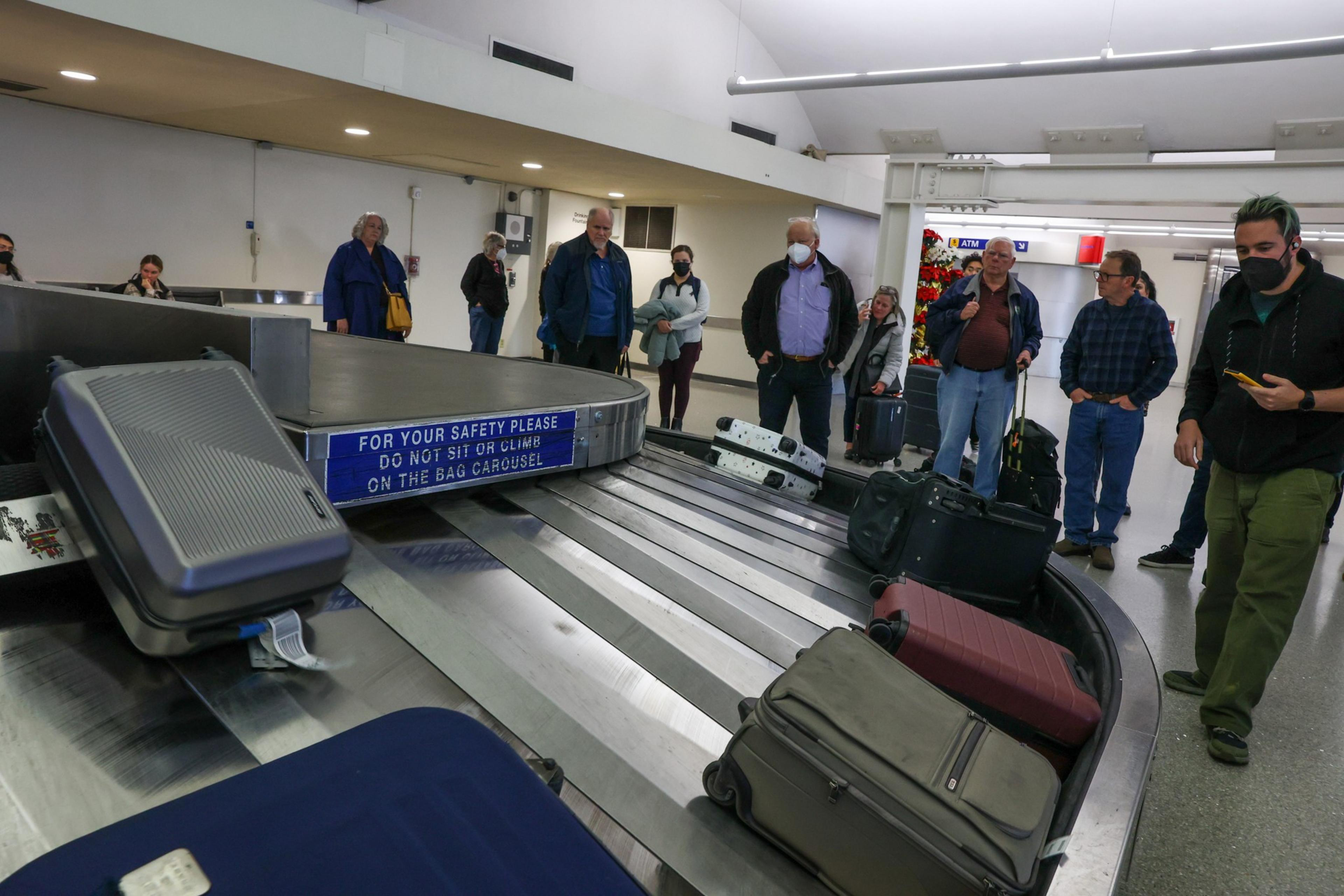 People stand around an airport baggage carousel, waiting for their luggage. Some wear masks, and several suitcases are in view. A safety sign is visible.
