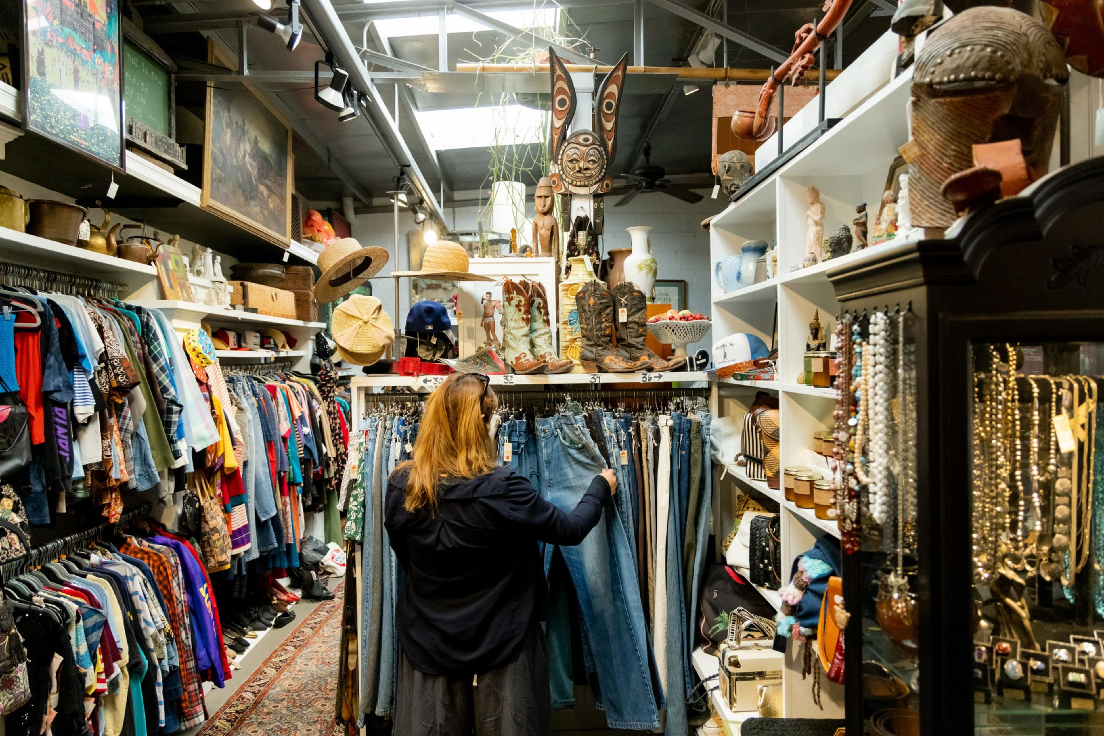 A woman browses denim jeans in a vibrant vintage store filled with colorful clothes, hats, boots, jewelry, and eclectic decorations on shelves and walls.