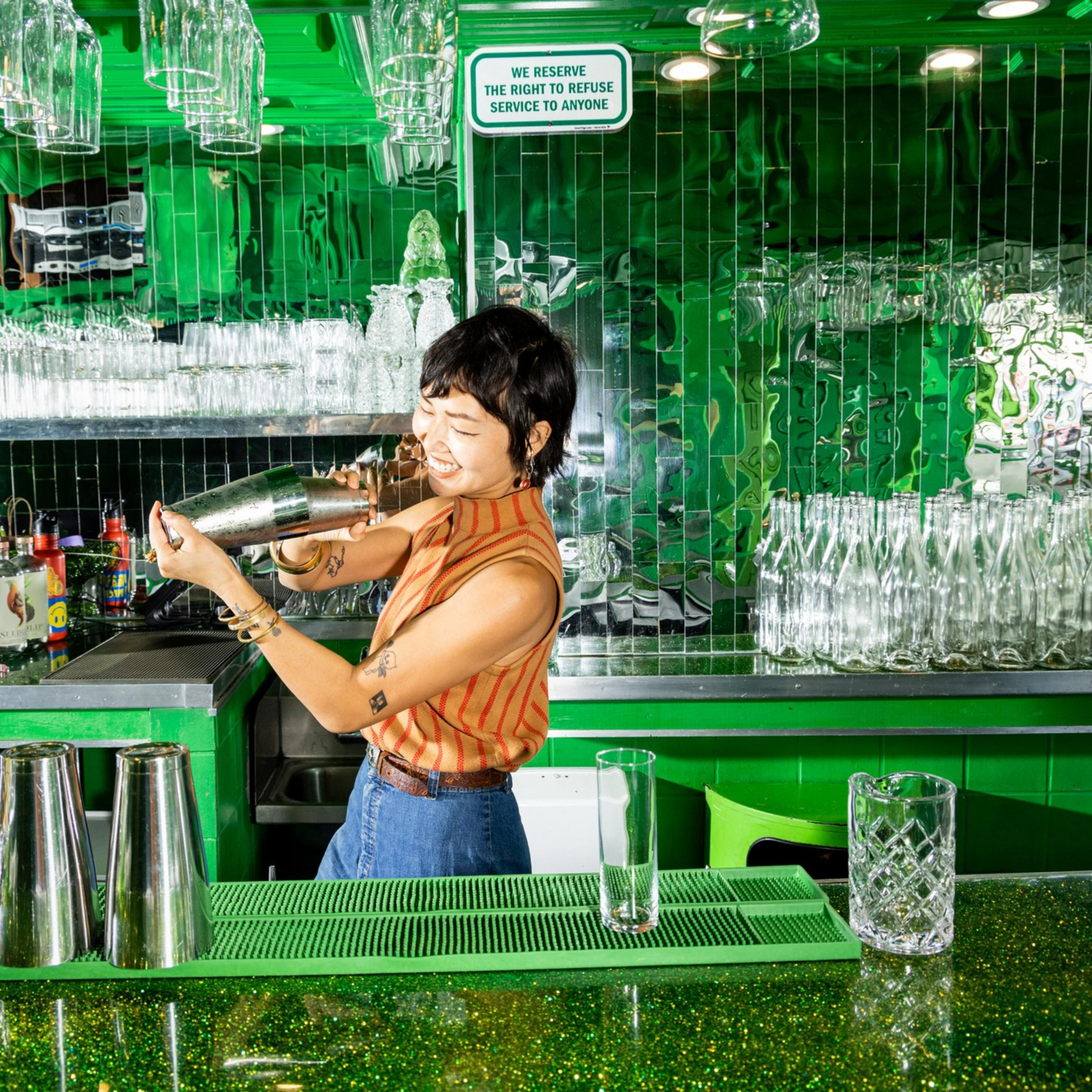 A bartender shakes a cocktail shaker behind a green-glittered bar with colorful straws, bottles, and glassware against a mirrored green wall.
