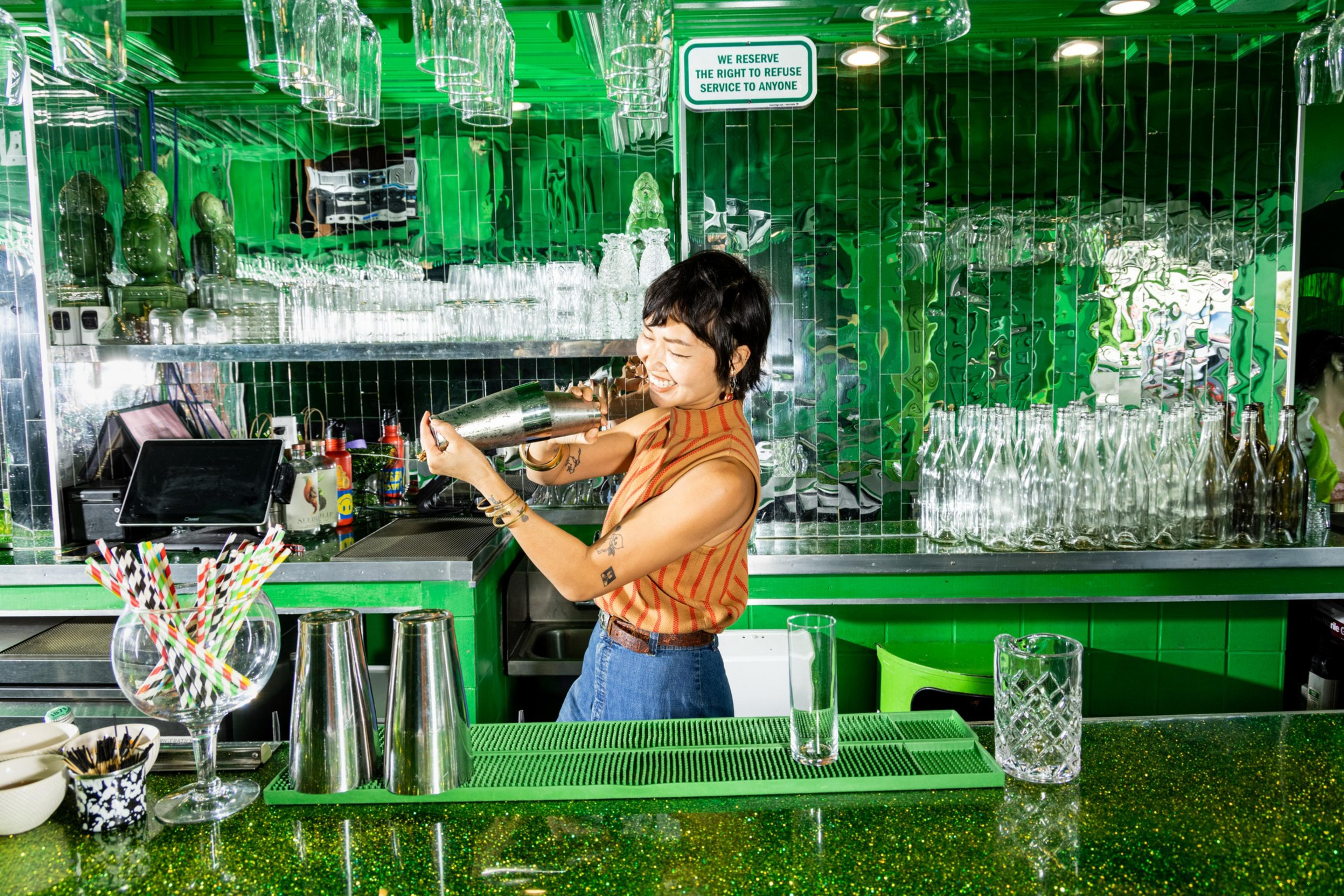A bartender shakes a cocktail shaker behind a green-glittered bar with colorful straws, bottles, and glassware against a mirrored green wall.