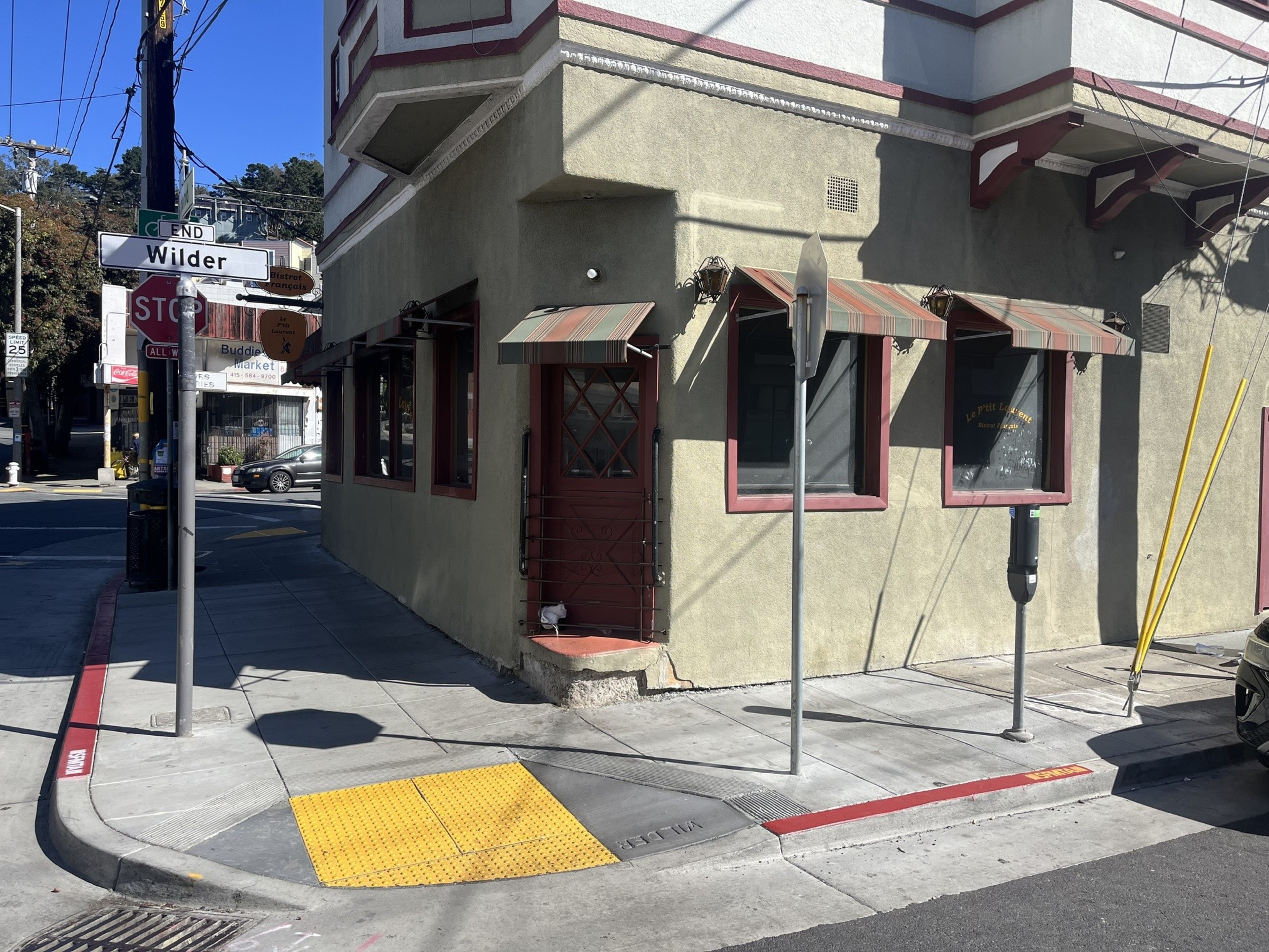 A street corner with a beige building featuring a red door, striped awnings, a parking meter, and street signs including an “End Wilder” sign.