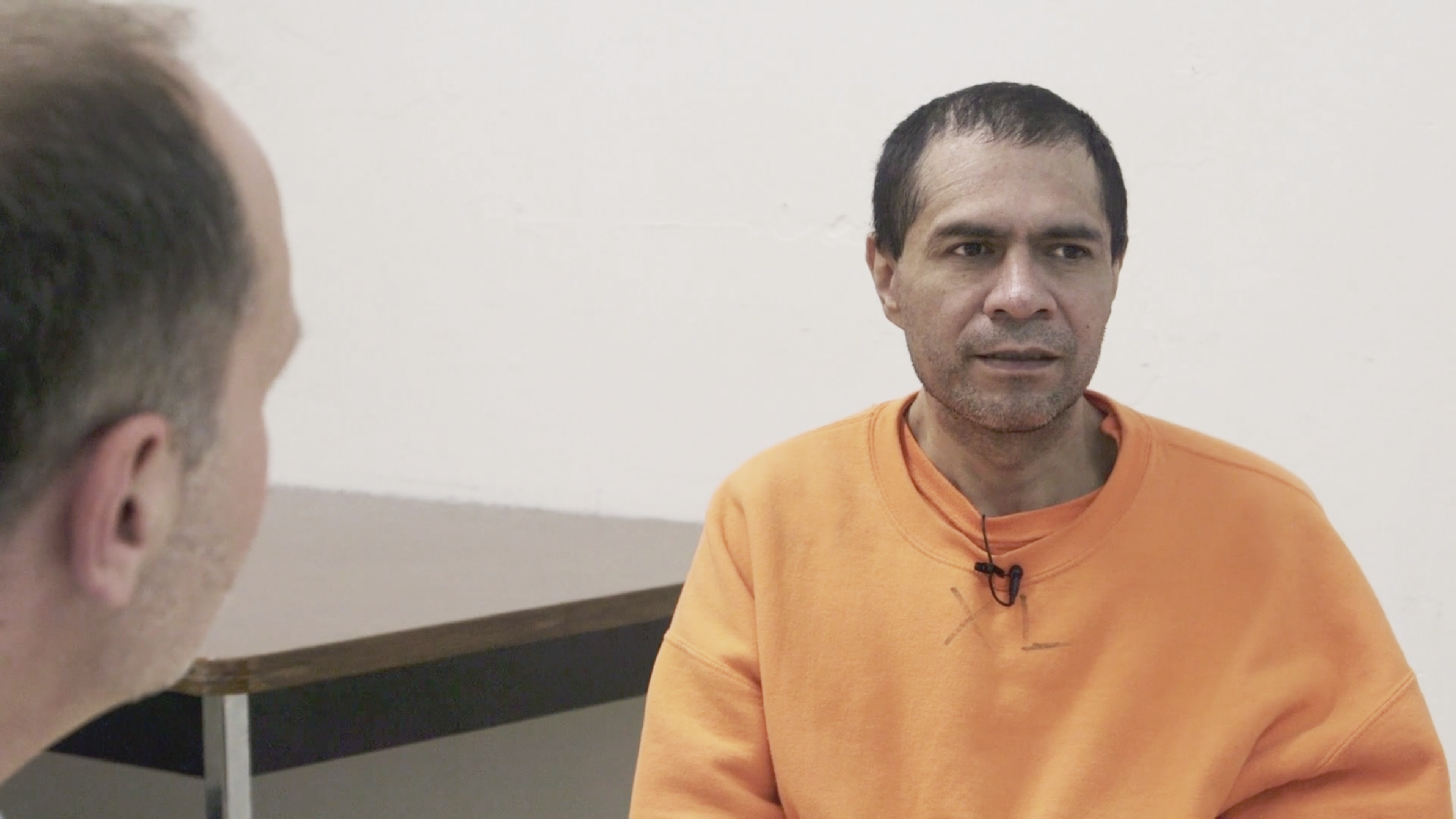 Two men are seated indoors; one facing away, the other in focus wearing a bright orange shirt, appearing engaged in conversation.