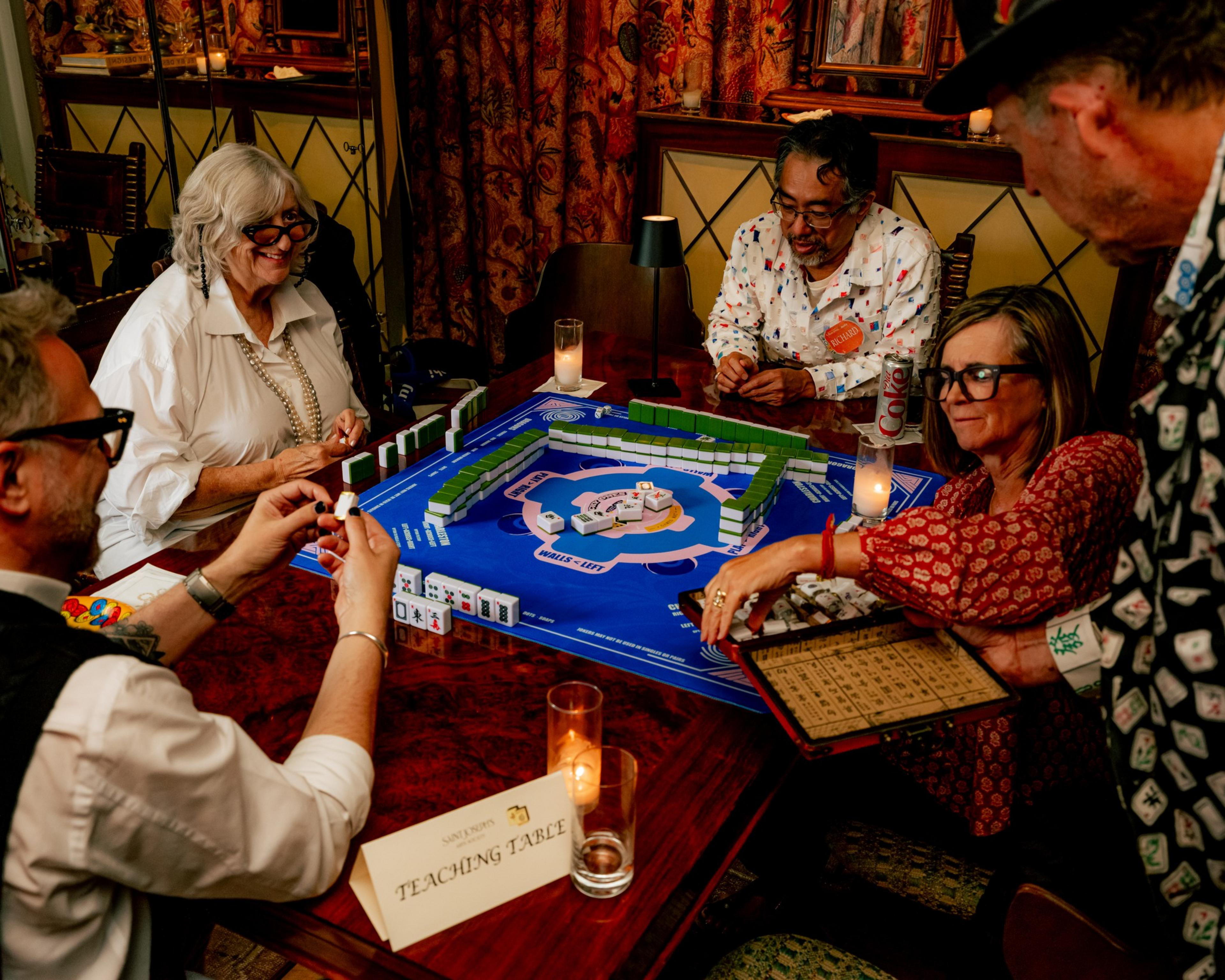 Five people with glasses gather around a table playing Mahjong with tiles on a blue mat labeled "TEACHING TABLE."