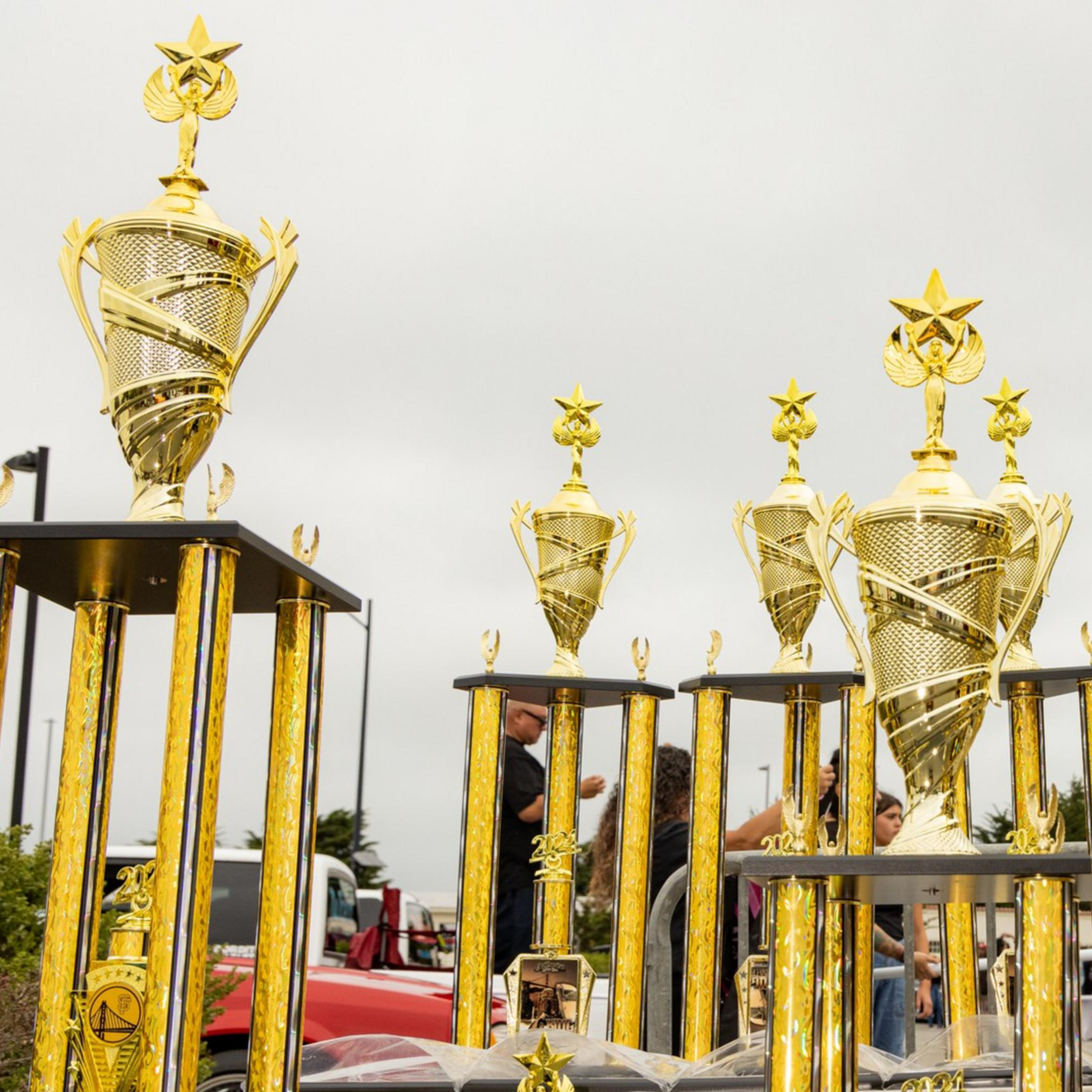 The image shows a variety of tall, golden trophies with intricate designs and star tops, displayed on tables outdoors with a cloudy sky in the background.
