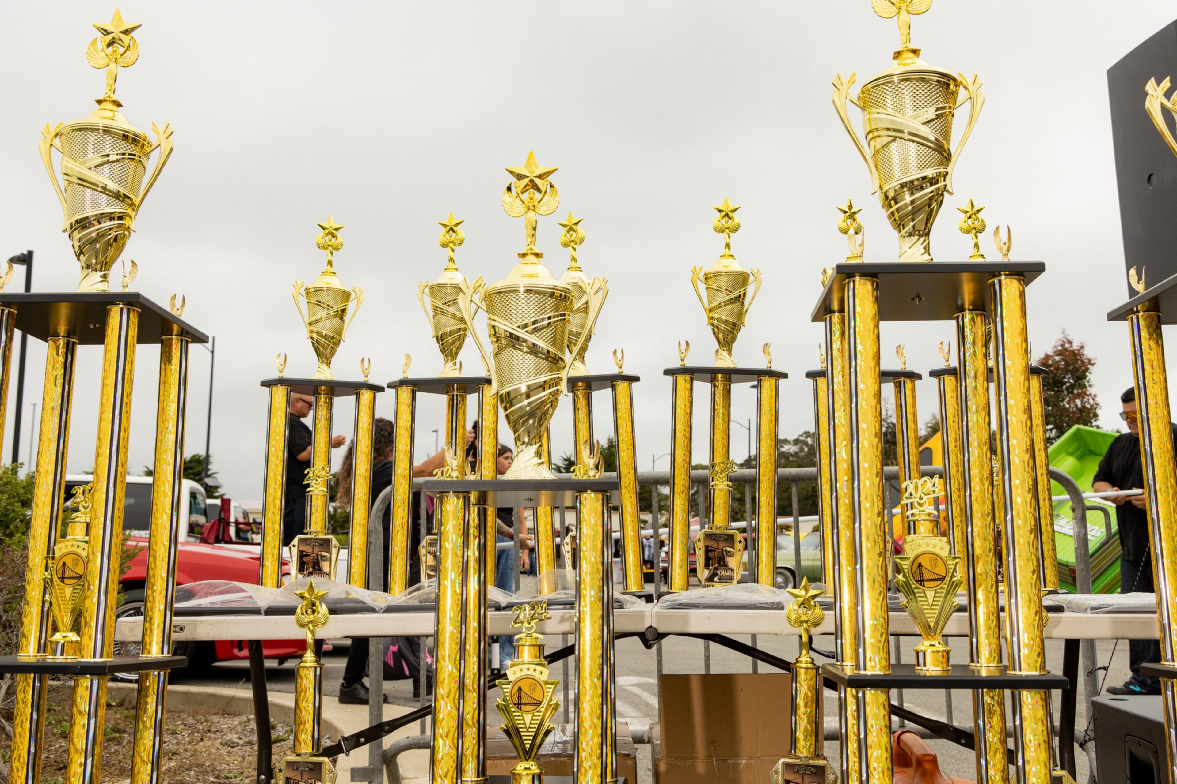 The image shows a variety of tall, golden trophies with intricate designs and star tops, displayed on tables outdoors with a cloudy sky in the background.
