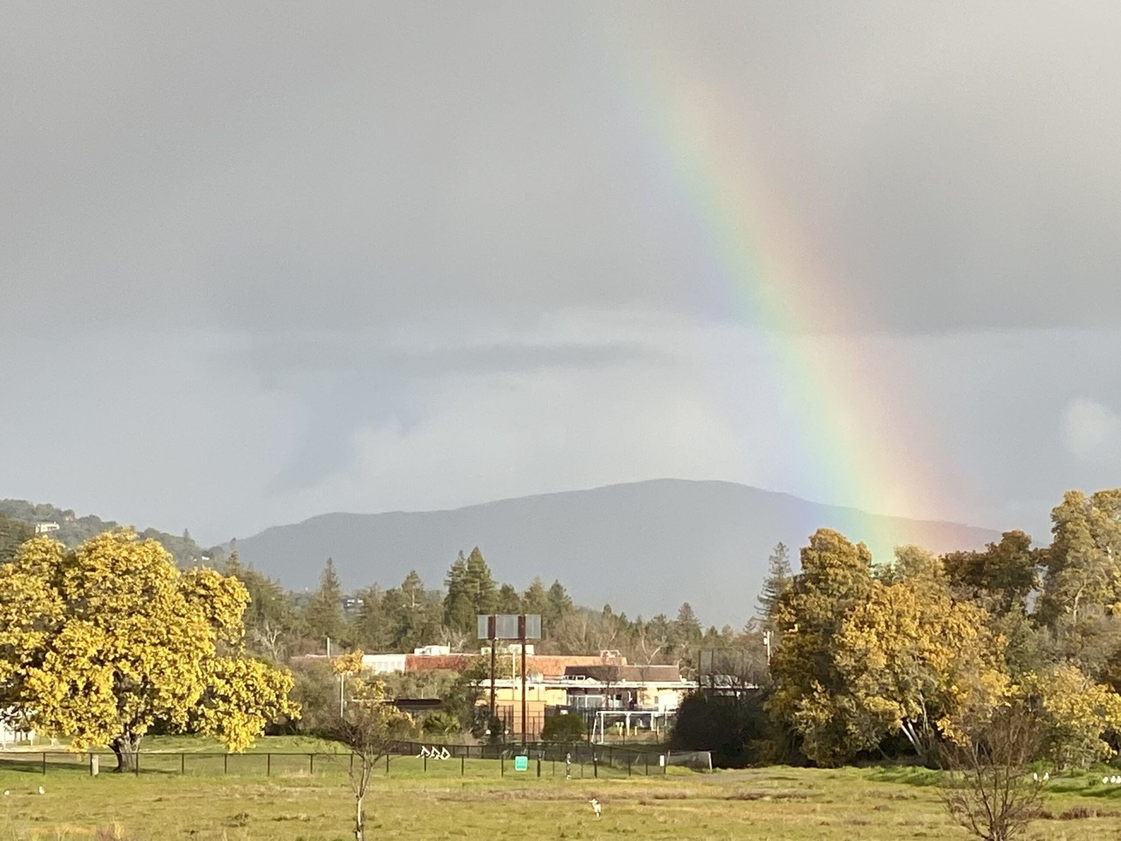 A vibrant rainbow arches over a distant hill, with buildings and trees in autumn hues beneath a cloudy sky.