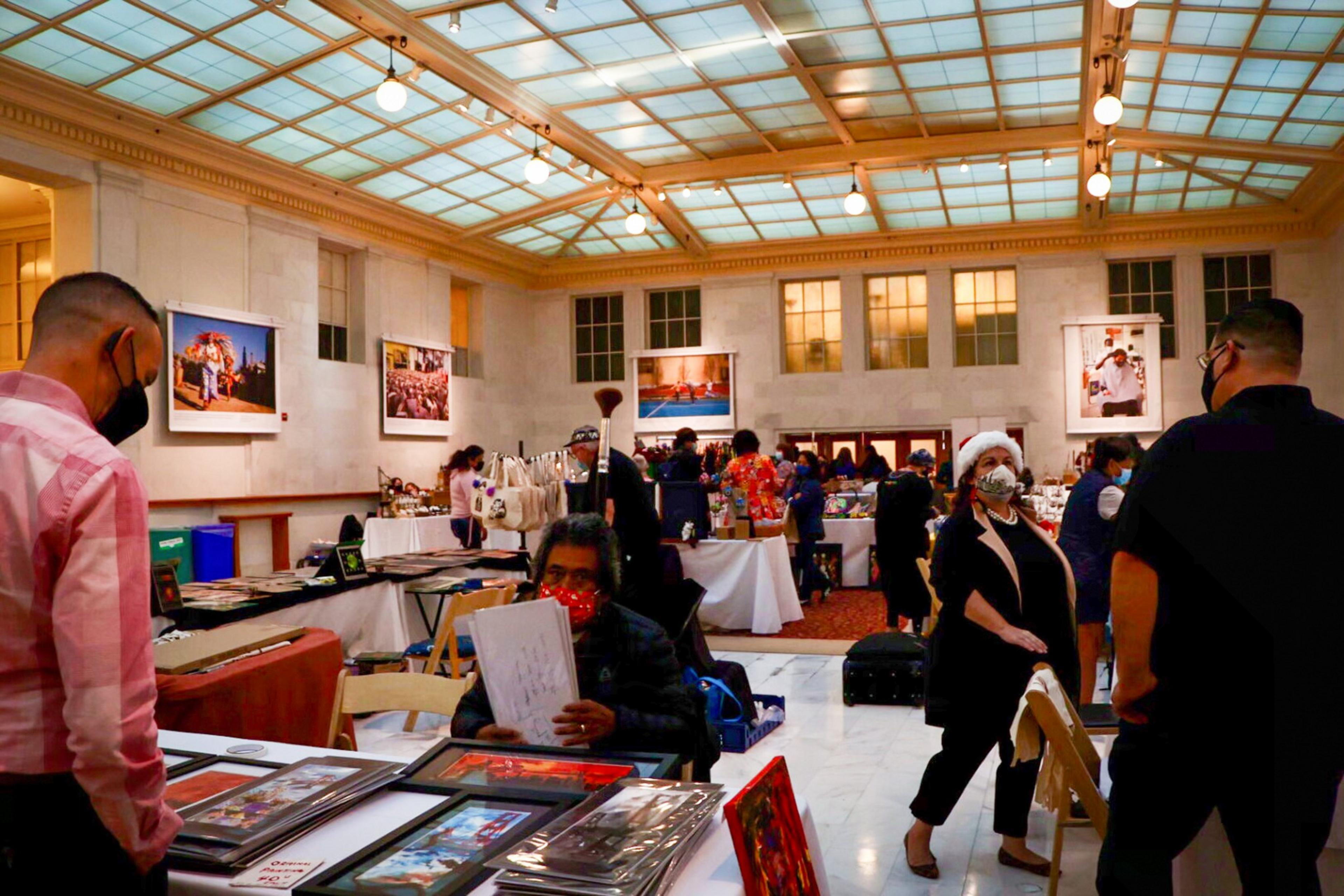 People wearing masks browse and sell artwork and crafts at tables inside a large, well-lit room with high ceilings and framed photos on the walls.