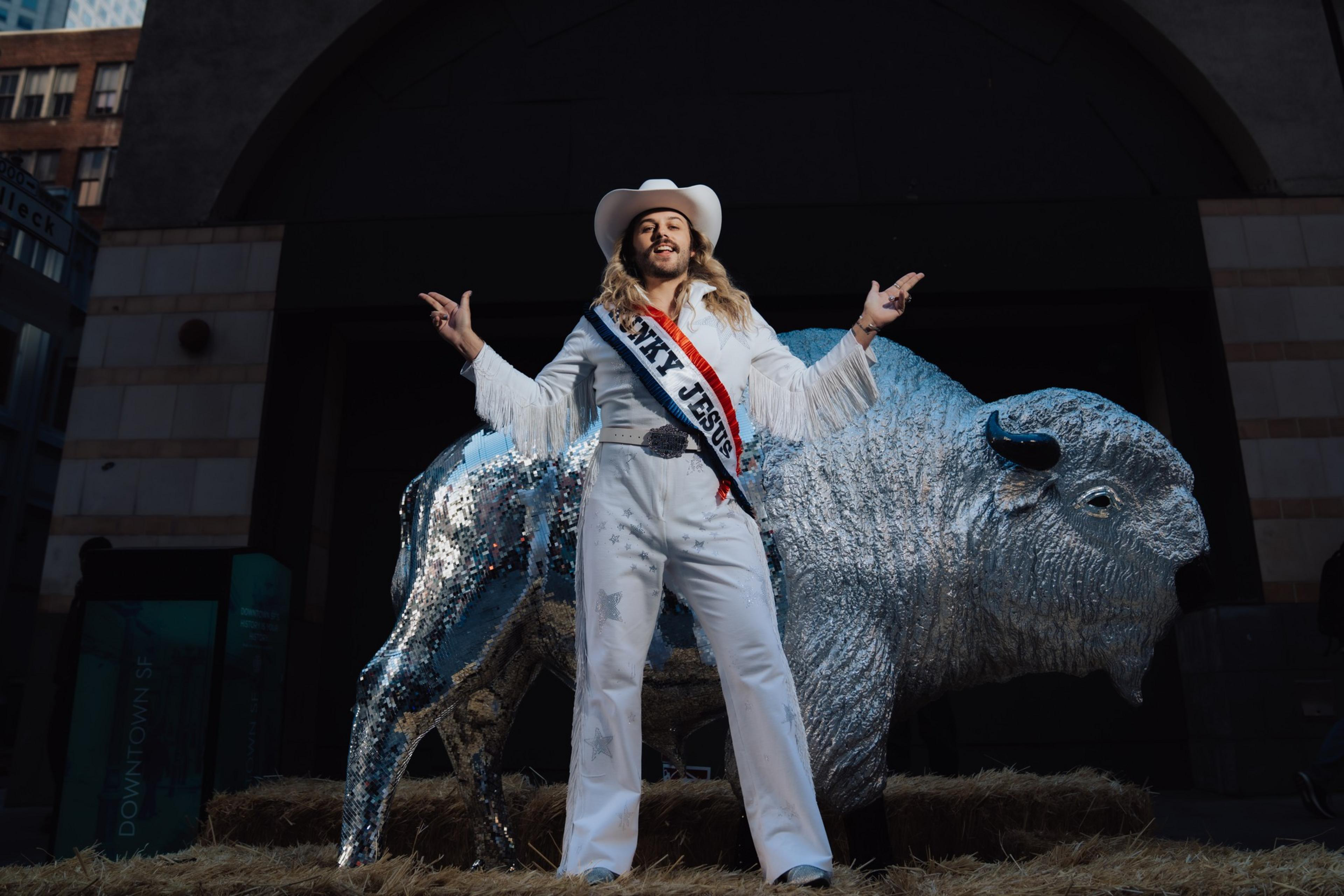 A person in a white outfit with fringe and a cowboy hat stands confidently in front of a shiny, mirrored bison statue, on a bed of hay.