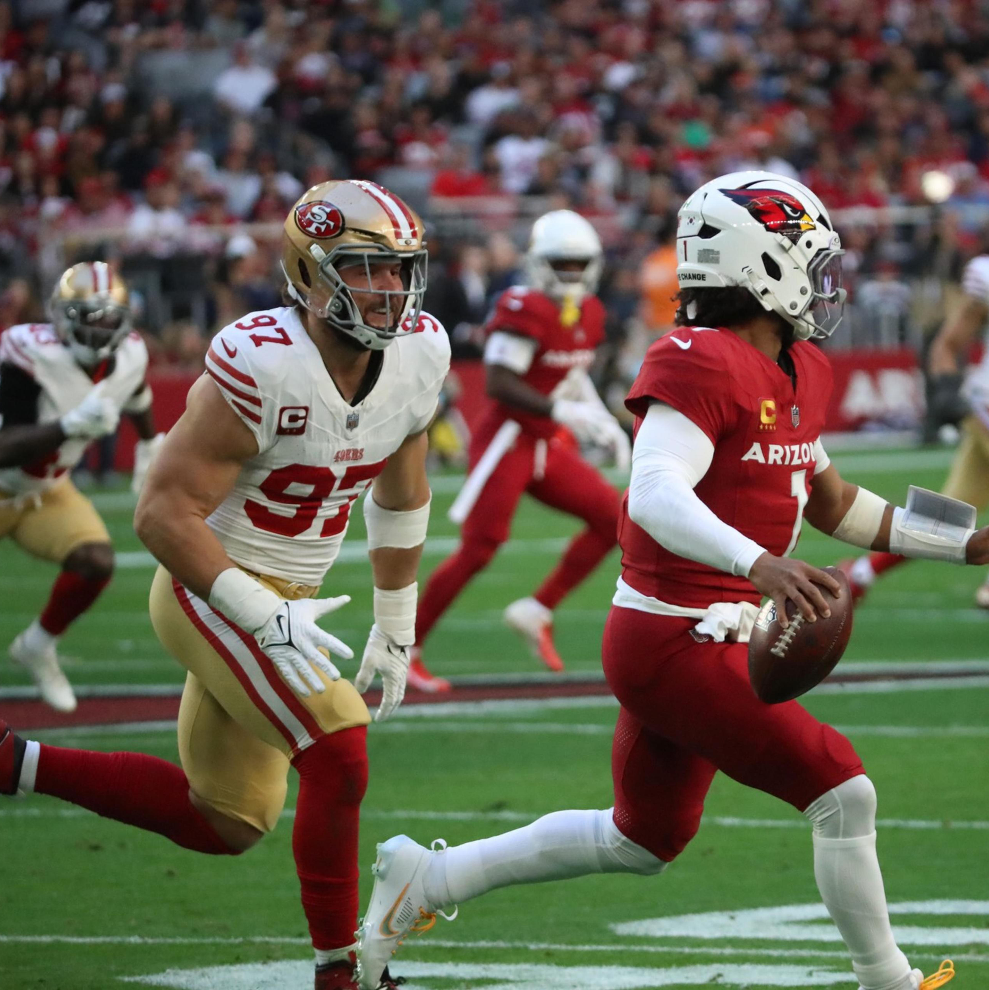 A quarterback in a red Arizona Cardinals uniform runs with the football as two San Francisco 49ers defenders chase him on a football field.