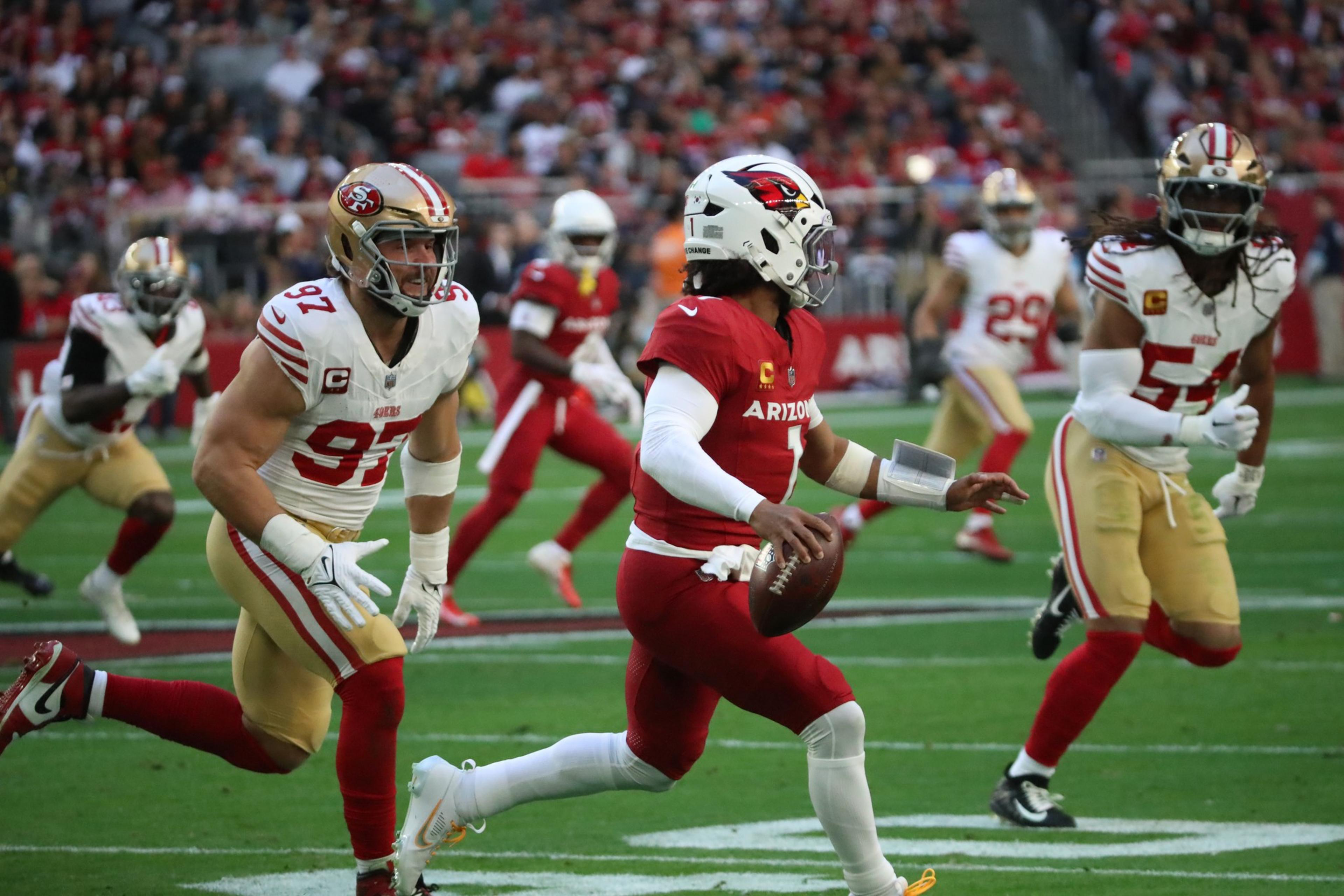 A quarterback in a red Arizona Cardinals uniform runs with the football as two San Francisco 49ers defenders chase him on a football field.