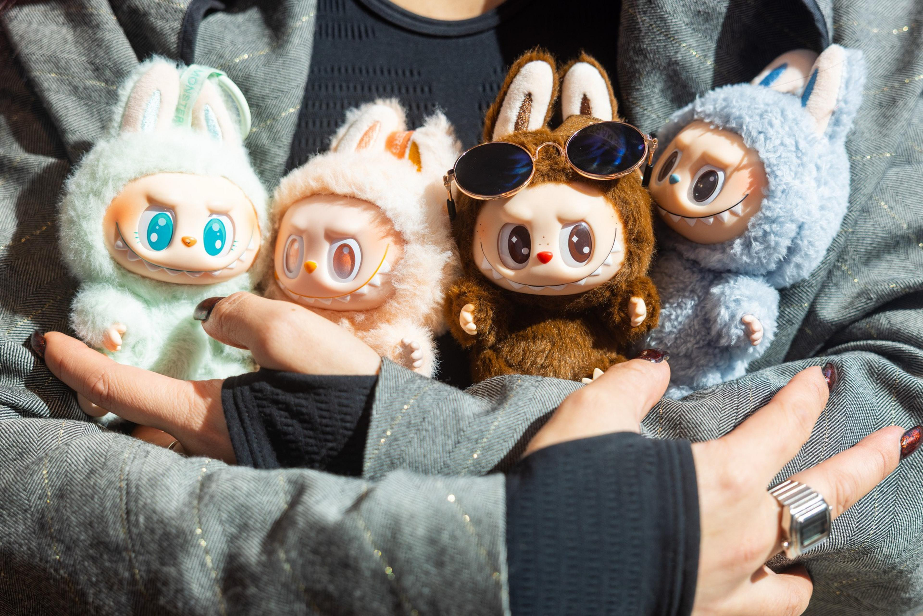 A person holds four plush toys in pastel and brown colors, each with big eyes and bunny ears. One toy wears sunglasses. The person’s nails are painted, and they wear rings.