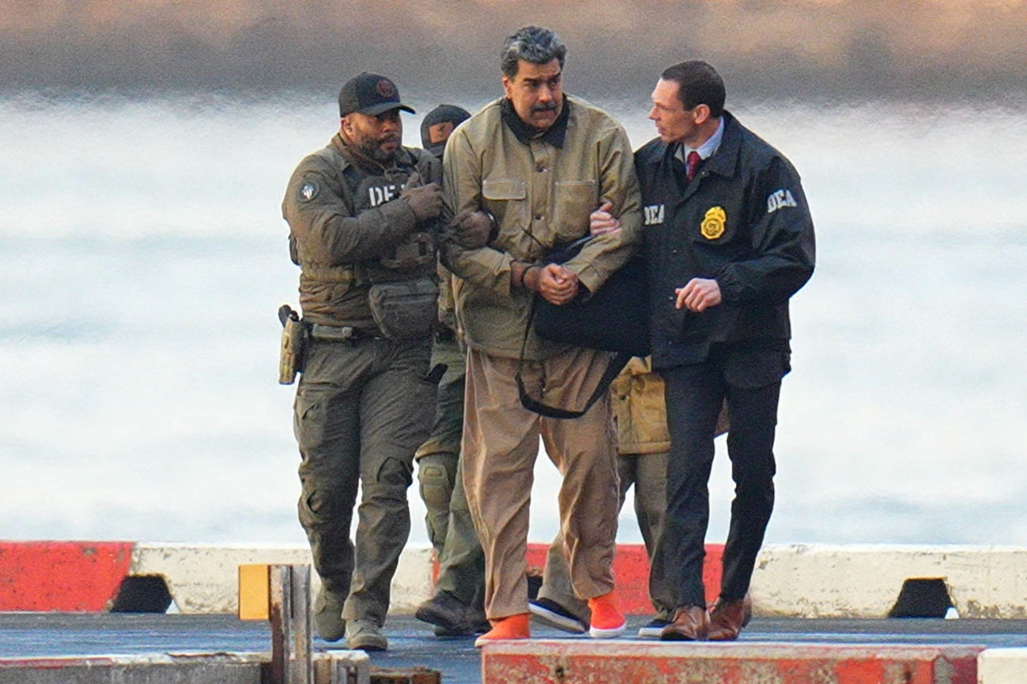 A man in a tan jacket and pants is being escorted by two officers in tactical gear and one in a DEA jacket near a waterfront.