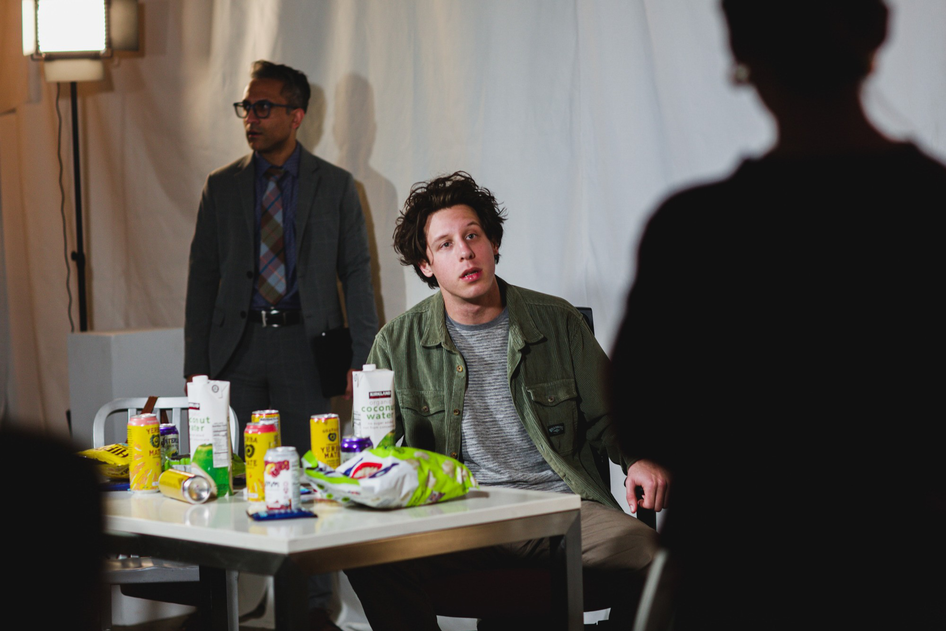 A man sits at a cluttered table with various drinks and snacks. Another man in a suit stands nearby, and a person is silhouetted in the foreground.