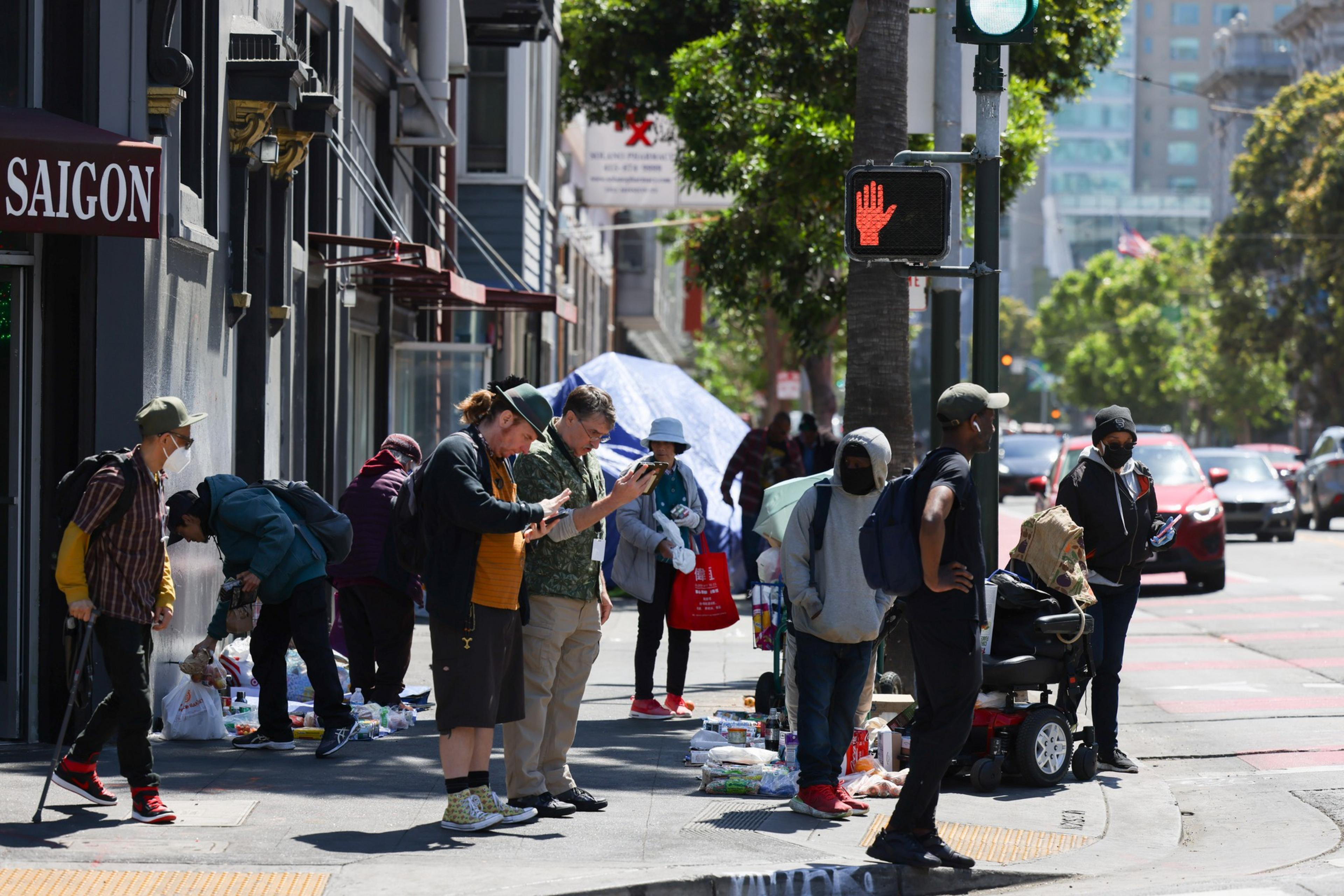 People gather on the corner of a city block, some waiting to cross the streets, others on their phones and some people selling merchandise that's on the sidewalk.