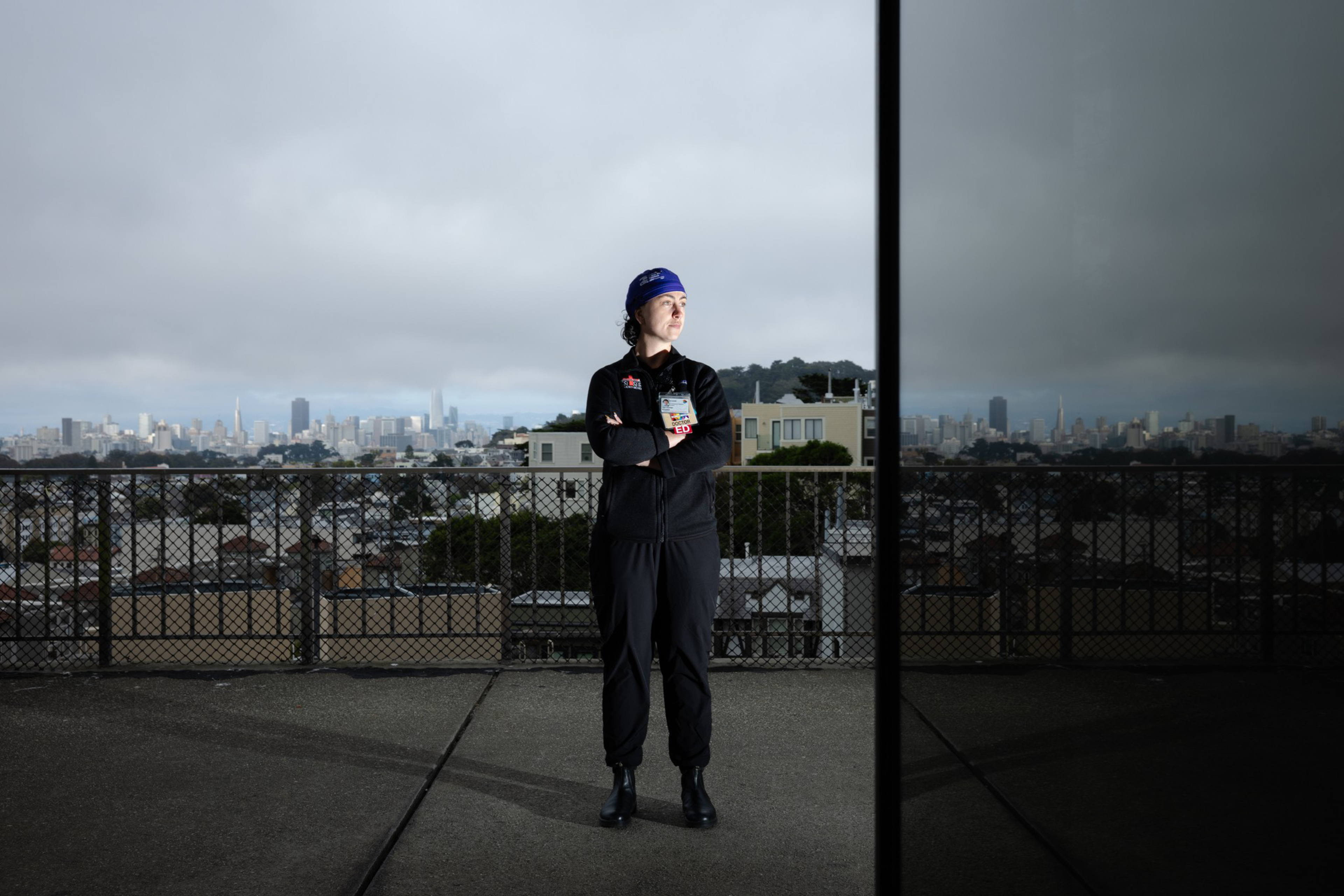 A person dressed in black with a blue headband stands with arms crossed on a rooftop, city skyline and cloudy sky visible behind them.