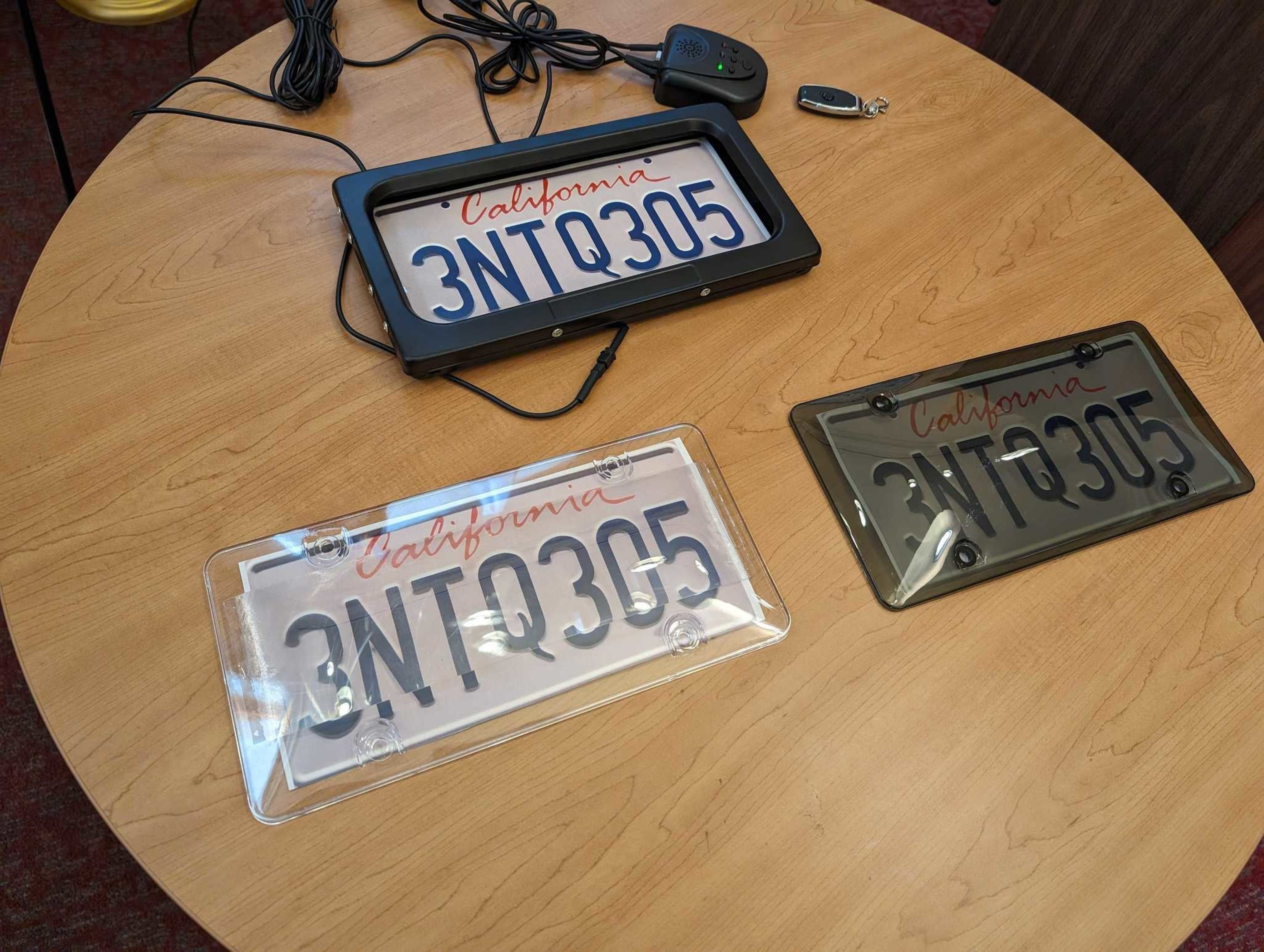 A circular wooden tabletop holds three versions of the same California license plate. One version is in an automatic device that responds to a key fob button, shuttering a sliding black door over the plate. Another version covers the plate with semitranspa