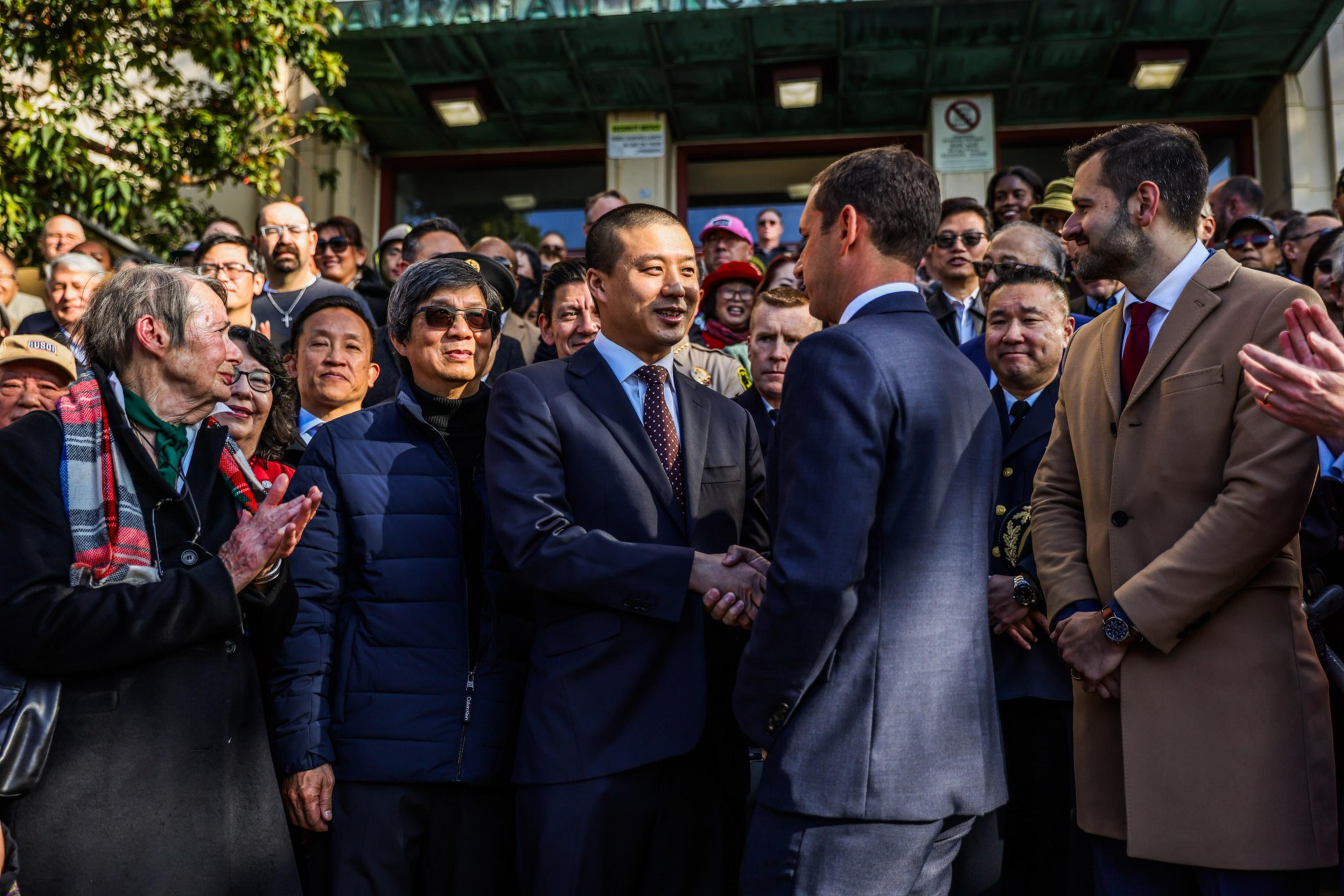 Two men in suits shake hands while a diverse crowd behind them watches and claps outside a building on a sunny day.