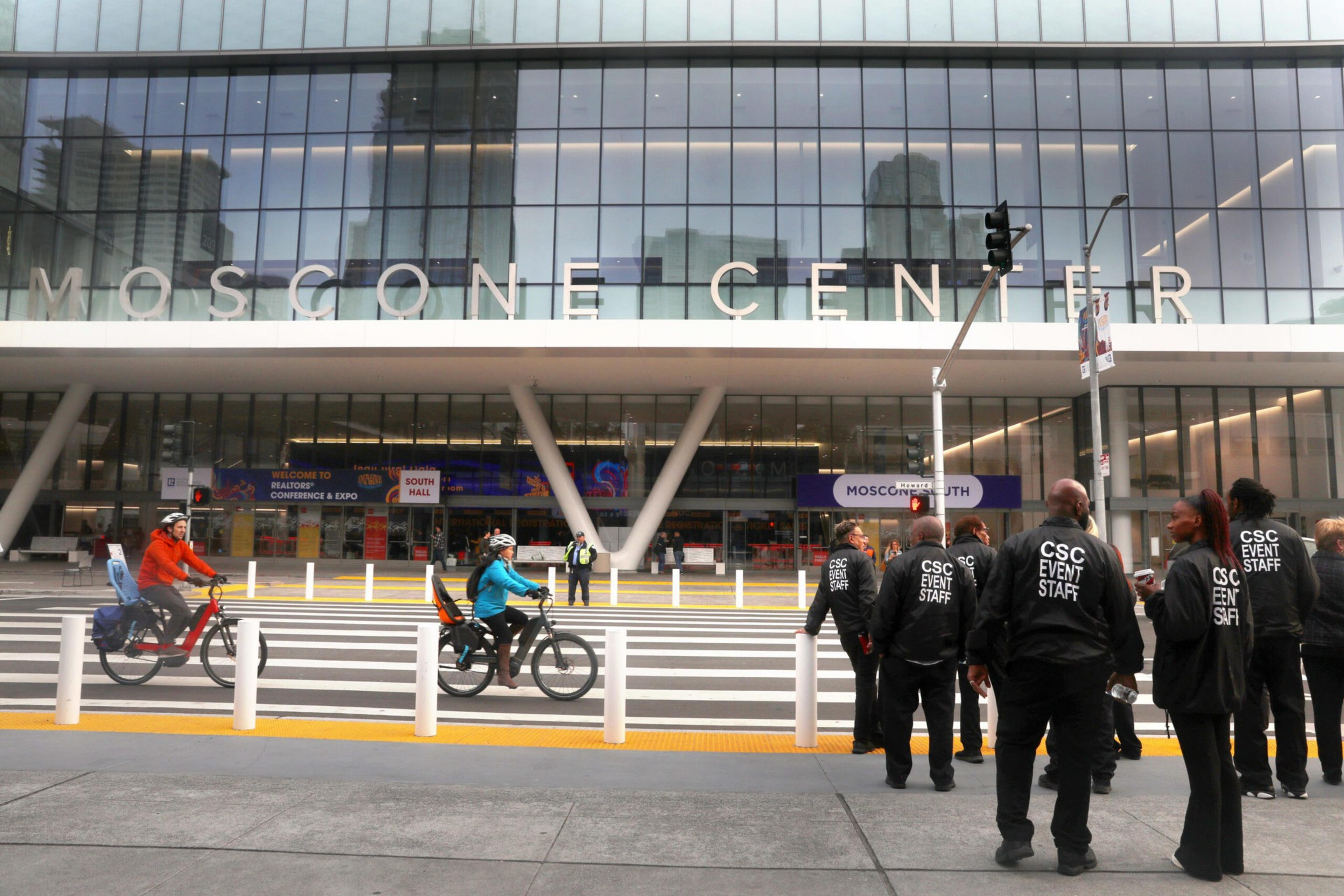 A group of event staff wearing black jackets stands outside Moscone Center while two people ride bikes on the street in front of the building.