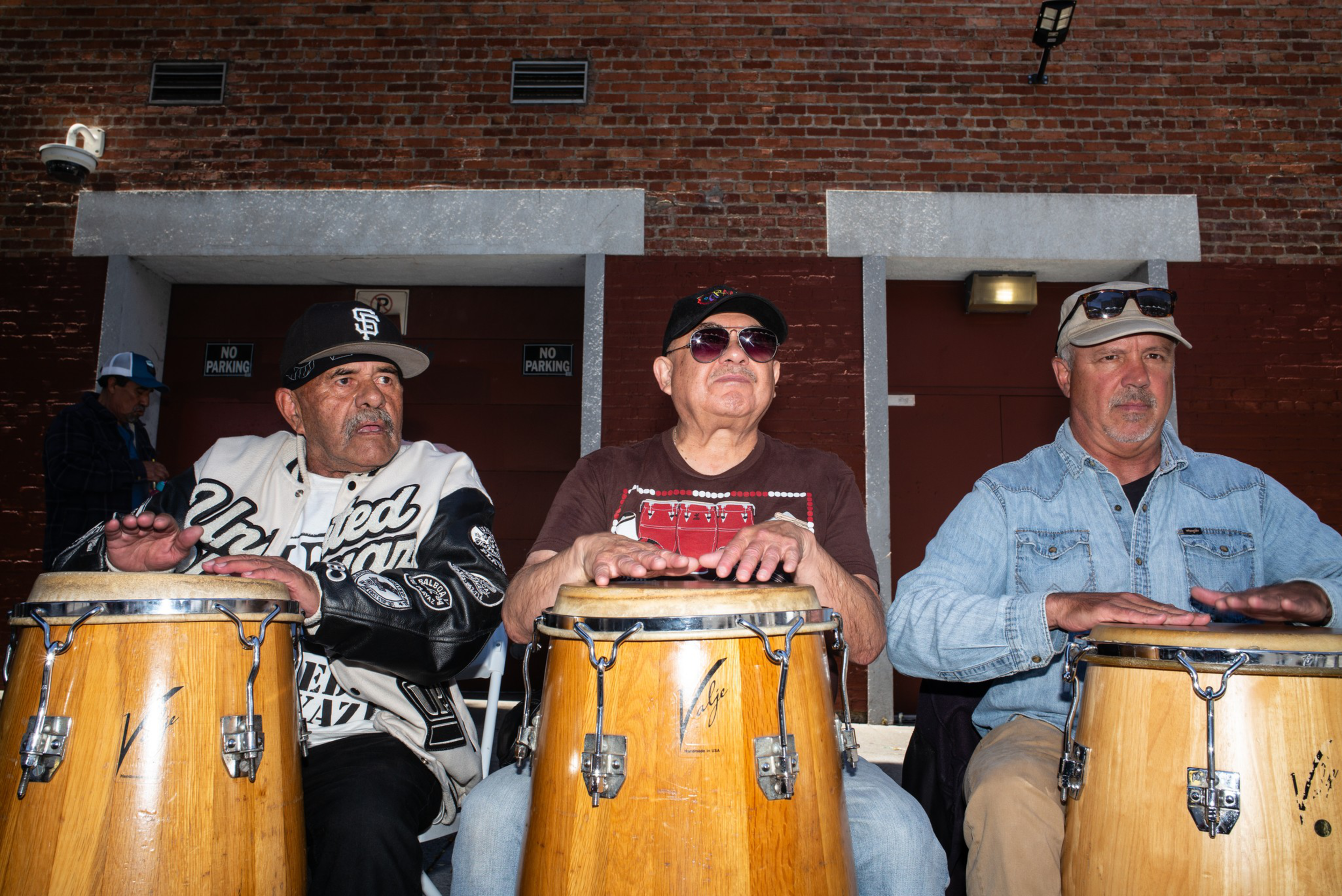 Three men sit side by side playing conga drums in front of a red brick wall, wearing casual clothes and hats under bright lighting.